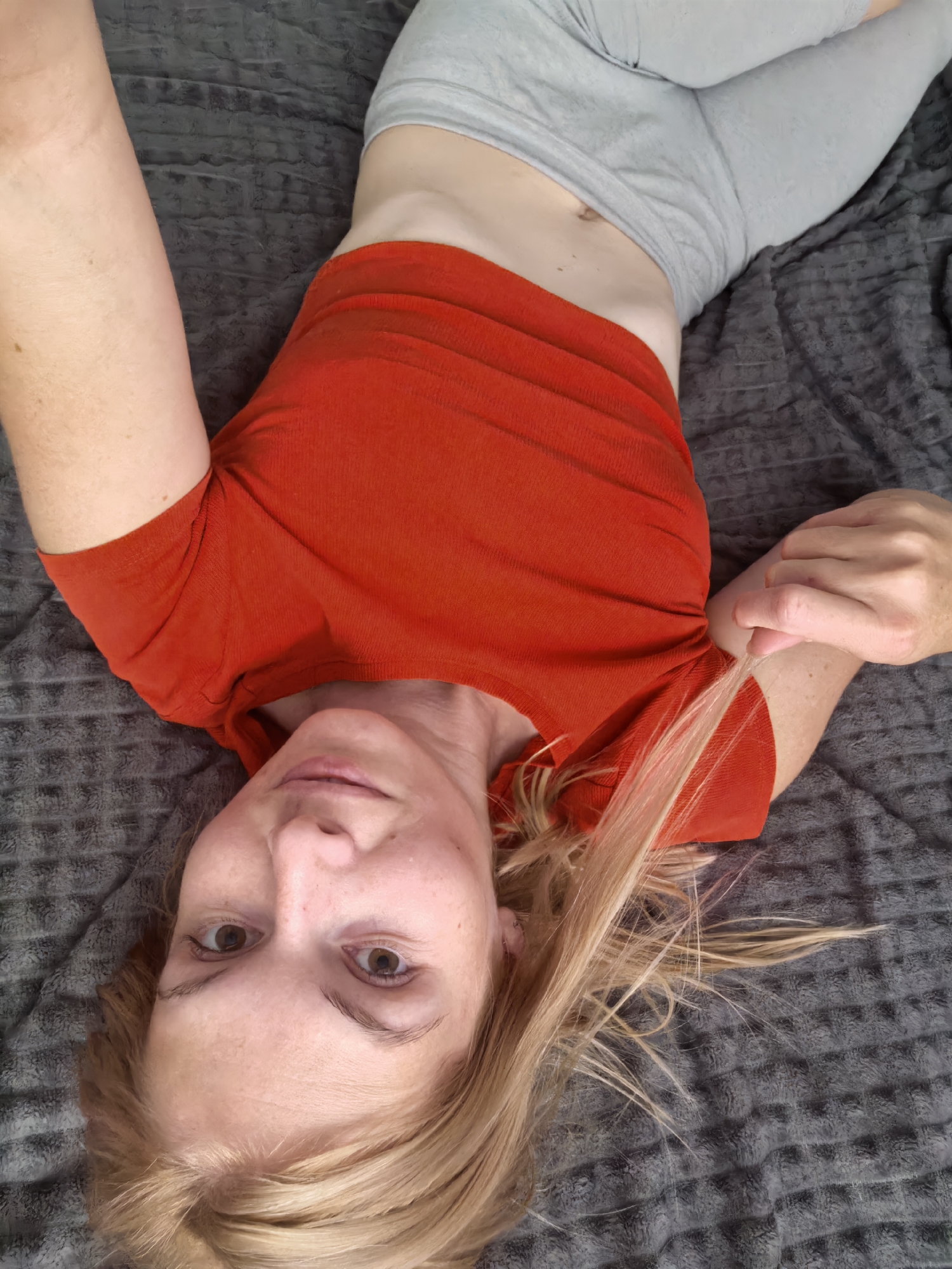 A woman with blonde hair, wearing a red t-shirt and grey shorts, is lying on a grey textured blanket. Her head is tilted to her left, and her right hand is pulling at the hem of her t-shirt. She is looking directly at the camera. The image was taken indoors. There is no specific information available about the location in the city of Sterlitamak, Russia.