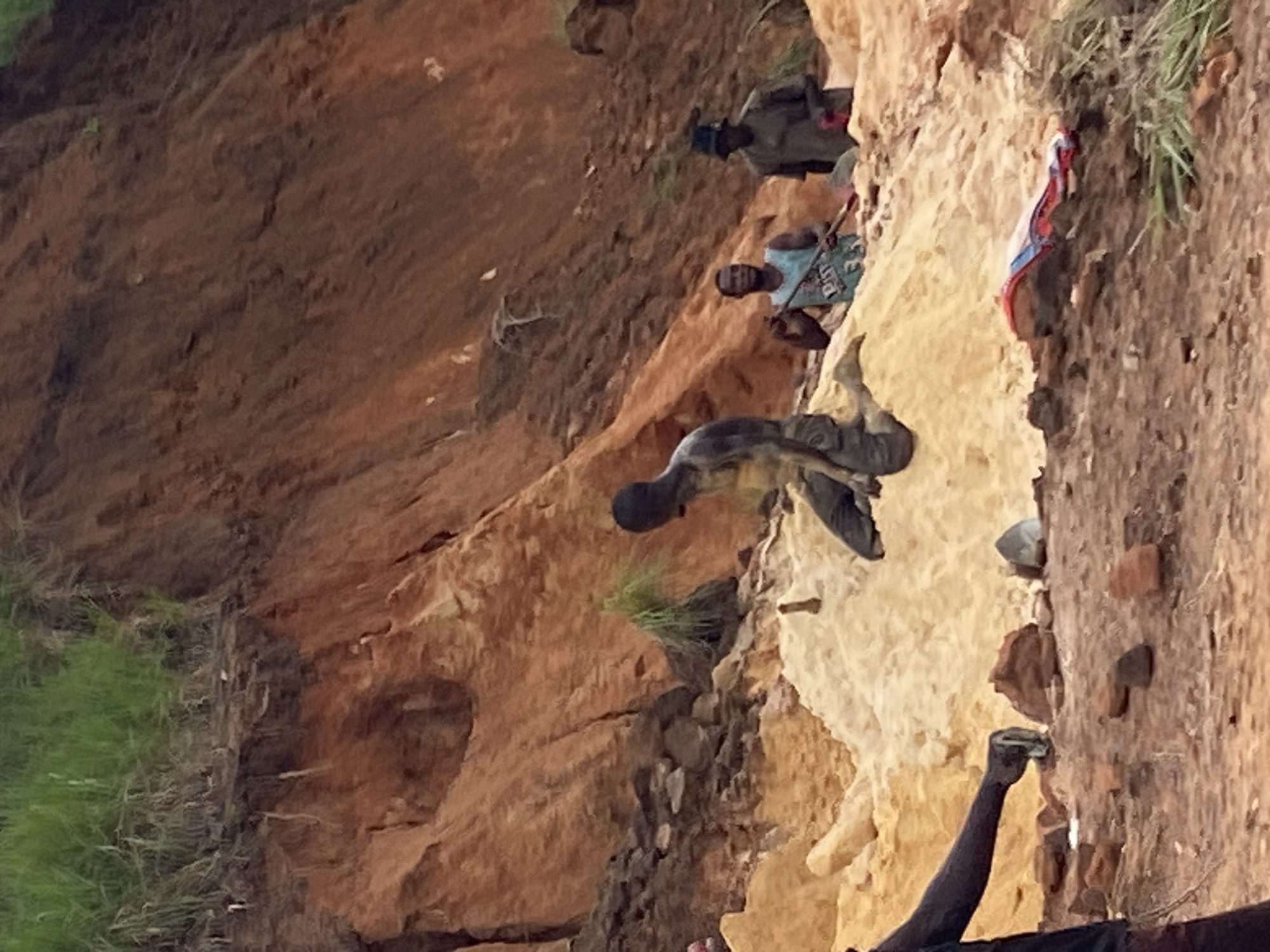 In the city of Ufu Esioni, Nigeria, a group of individuals is engaged in mining activities. The scene is characterized by a steep, eroded embankment of reddish-brown soil, topped with sparse green vegetation. In the foreground, a man is kneeling on a mound of light-colored sand, seemingly working with his hands. To his right, another man stands wearing a blue and white patterned tank top and holding a long stick. Further to the right, a third man is visible, wearing a hat and a dark shirt. The ground is covered with dirt, stones, and piles of sand. A red and blue object, possibly a piece of fabric or clothing, is lying on the ground in the lower right portion of the image. The overall environment appears to be a raw, uncultivated mining site.