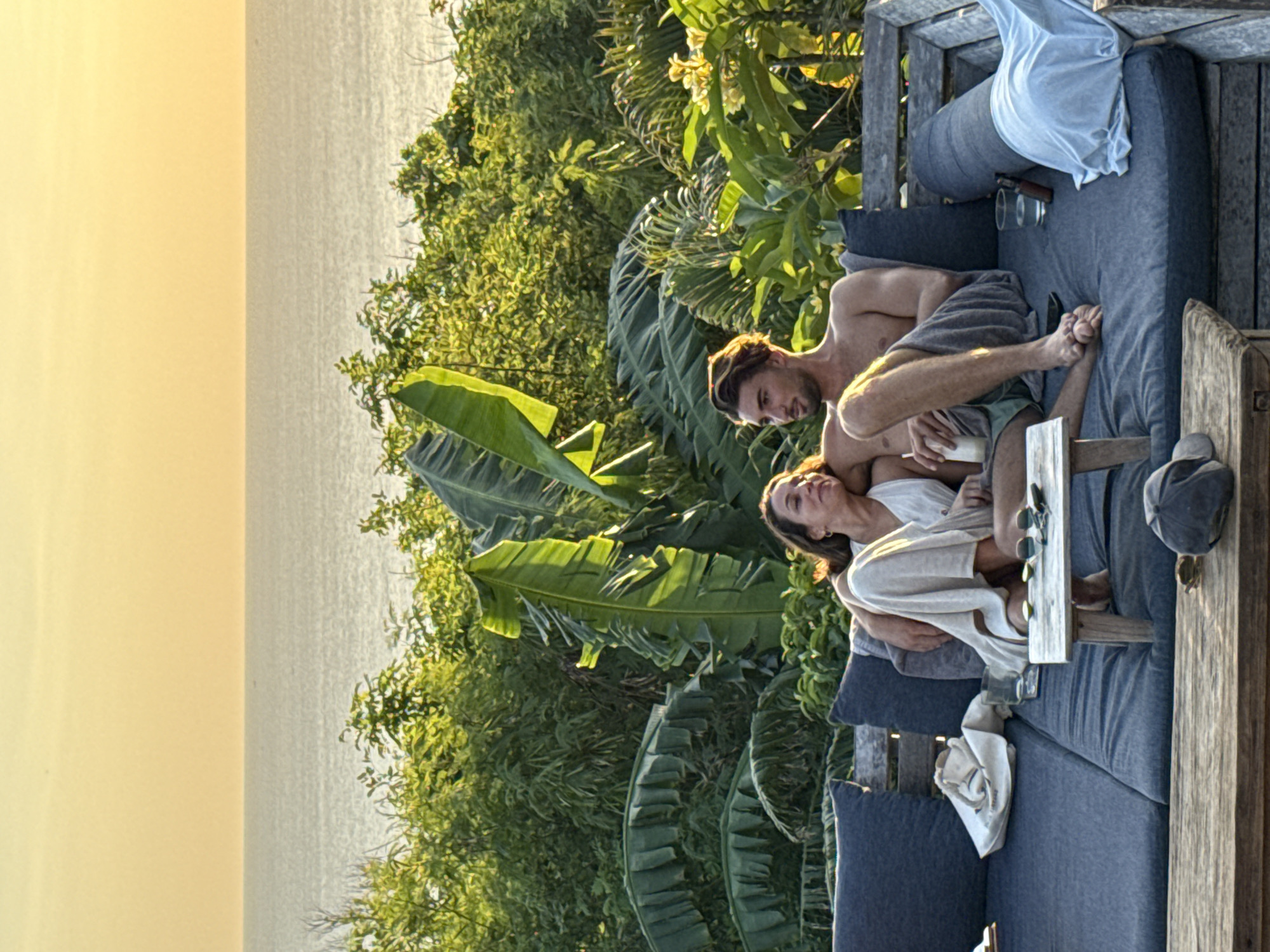 A man and a woman are embracing on a cushioned outdoor lounge sofa with glasses, sunglasses, and a hat on a small table in front of them. The background includes lush tropical vegetation and a view of the ocean in Kuta Selatan, Indonesia. The lighting suggests it is either sunrise or sunset.