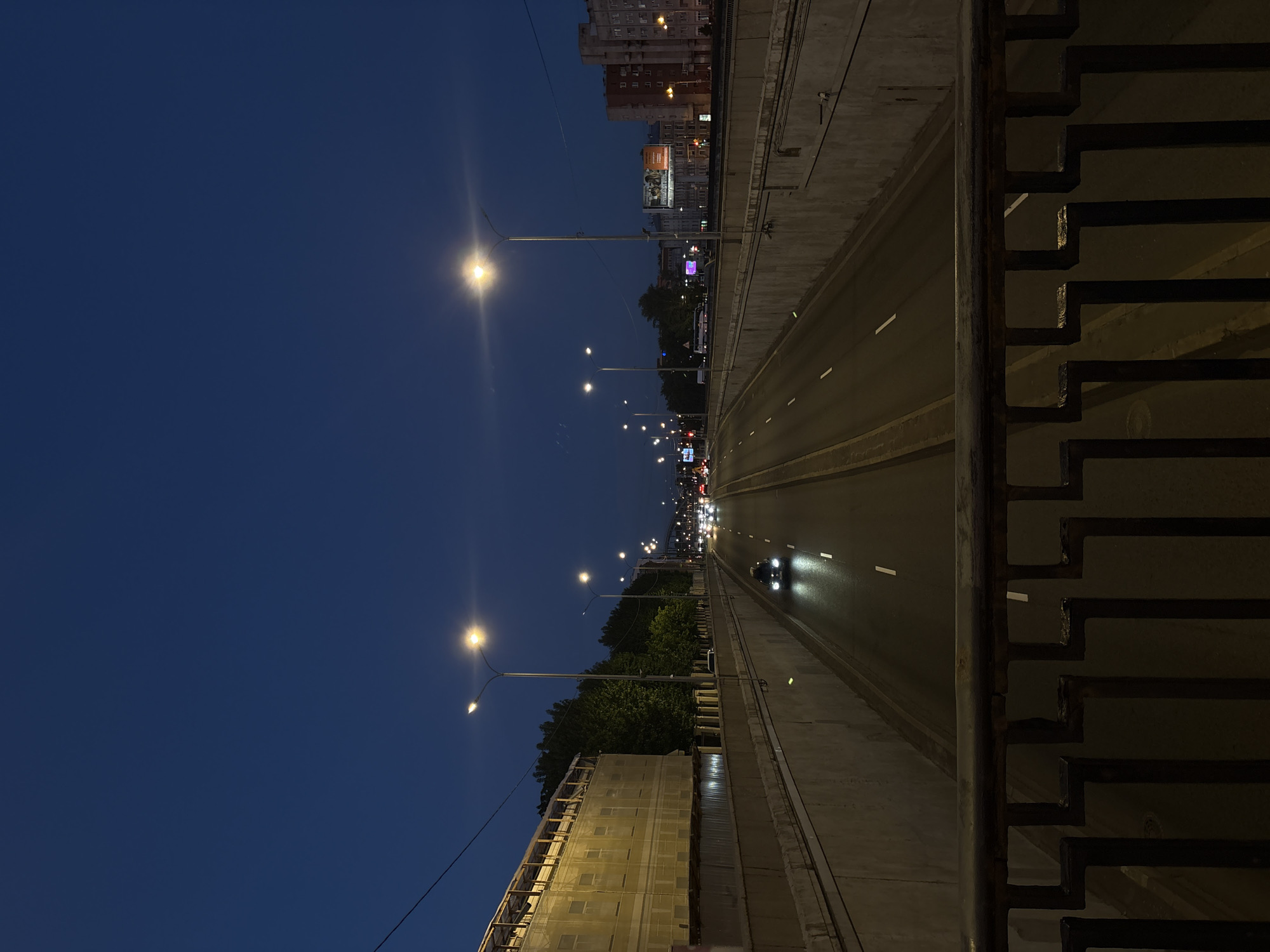 The image depicts a view of a highway interchange in Saint Petersburg, Russia, at dusk or night. The sky is a deep, dark blue, indicating the transition from daylight to darkness. Streetlights line the highway, casting an artificial glow on the roadway. Several vehicles are visible on the highway, their headlights and taillights creating streaks of light. The highway is flanked by concrete retaining walls, suggesting it might be a below-grade or elevated road. On the left side of the highway, there is a building under construction, partially covered with netting. Trees are visible behind the retaining wall, adding a touch of nature to the urban landscape. On the right side, buildings, some appearing to be apartment blocks, are visible in the distance, illuminated by streetlights and internal lighting. The foreground features a dark, metal railing, suggesting the image was taken from a bridge or an overpass overlooking the highway. The overall impression is one of a bustling urban environment at night, with the flow of traffic continuing despite the low light conditions.