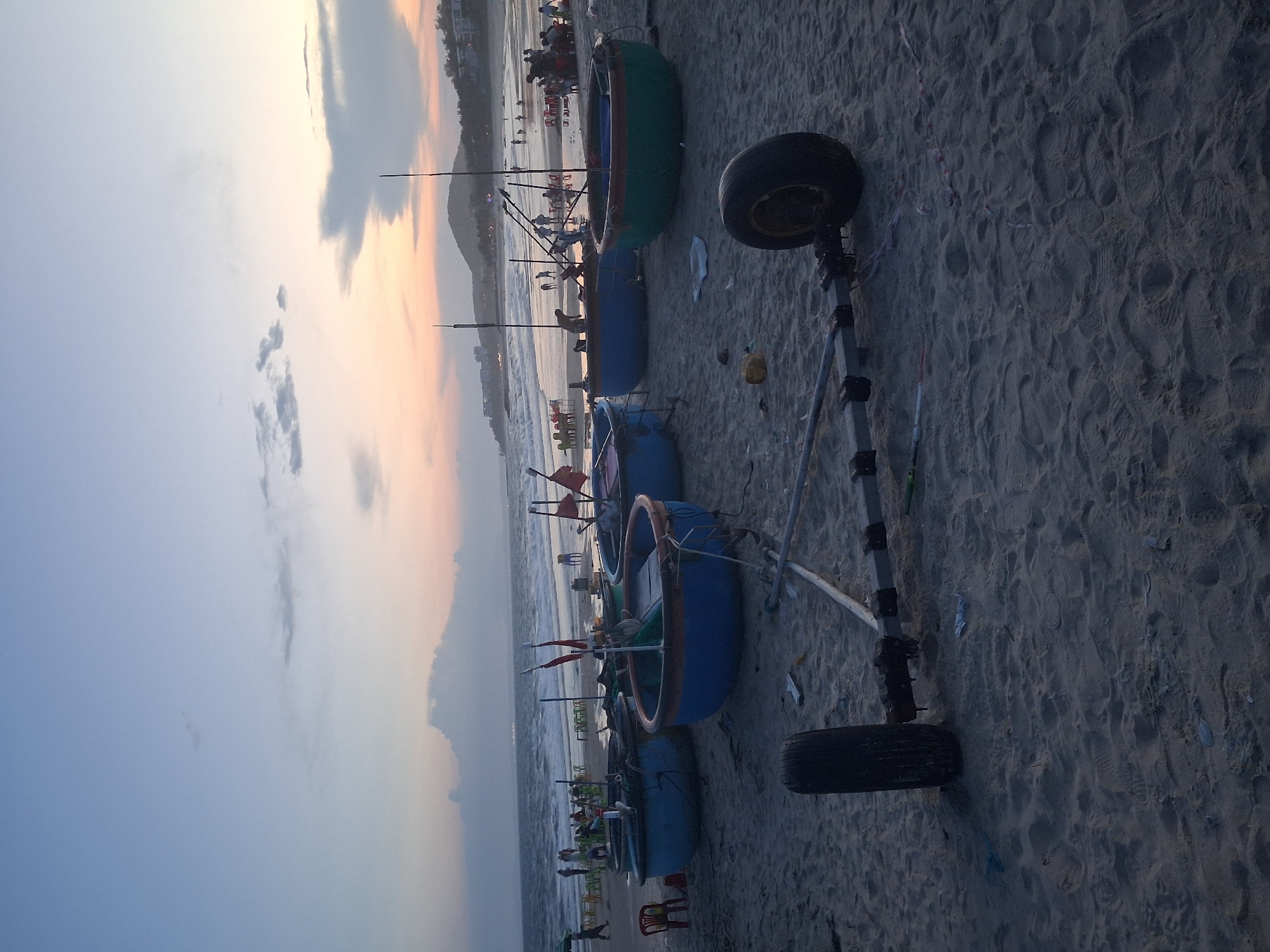 This media file depicts a beach scene at Phu Thuy Ward, Vietnam, during what appears to be sunset or sunrise due to the soft, warm light on the horizon and the overcast sky. The main subjects are several traditional Vietnamese basket boats, also known as coracles, lined up on the sandy beach. These boats are round and brightly colored, with one prominent green and blue boat in the foreground, and several blue ones further back.
A makeshift trailer with two wheels, likely used to move the boats, is positioned on the sand. One of the wheels is a black tire on a metal frame. The beach is covered in sand, showing numerous footprints, suggesting it's a public area. In the background, the ocean waves are visible, and beyond that, a coastline with buildings and trees can be seen, indicating a developed area near the beach. There are also some scattered plastic debris on the sand.
While the image is somewhat distant, small groups of people can be observed in the background near the water and along the beach, some appearing to be sitting on chairs. The overall atmosphere is tranquil, with the soft light creating a peaceful mood. The time of day is either dawn or dusk, given the sky's coloration. There is no visible text in the image.