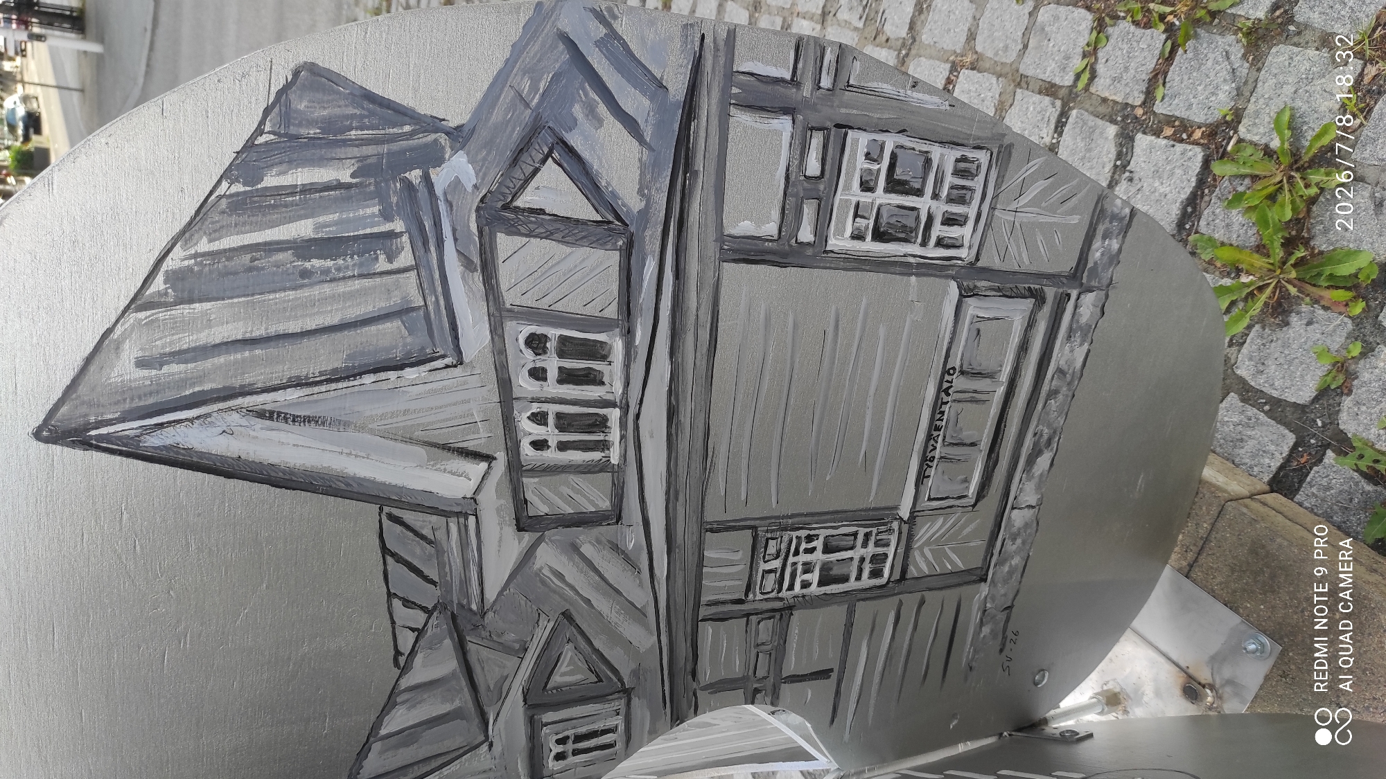 The image is a close-up, eye-level shot of a circular, metallic art installation painted in grayscale. The artwork depicts a building with a distinctive, steeply pitched roof and multiple windows, rendered in a stylized, somewhat abstract manner. The paint strokes are visible, giving the artwork a textured appearance.
The metallic surface of the art piece appears to be mounted on a metal pole, which is bolted to a concrete base. The installation is situated outdoors, with a cobblestone ground visible in the foreground and blurred background elements suggesting a street scene with vehicles and buildings.
Text is visible on the artwork: "TYÖVÄENTALO" is painted horizontally across a section of the building facade, and "SU-26" is inscribed below it. The red watermark "REDMI NOTE 9 PRO AI QUAD CAMERA" and the timestamp "2026/7/8 18:32" are overlaid in the lower portion of the image.
This artwork is located on the exterior of the Workers' House (Työväentalo) in Iisalmi, Finland. The building depicted in the artwork is likely the Iisalmi Workers' House itself, and the "SU-26" could refer to an address, a specific section, or a date related to the building or the artwork. The scene captures a detail of public art within the urban environment of Iisalmi.