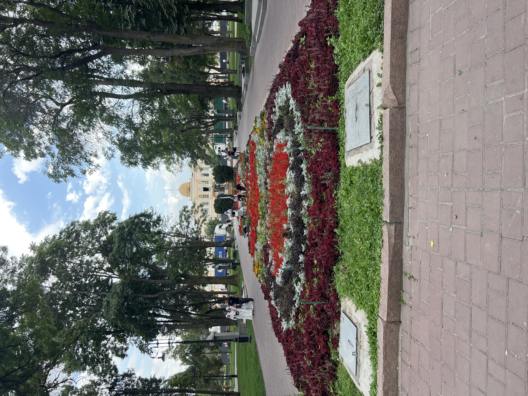 In Bishkek City, Kyrgyzstan, a wide, tree-lined pathway leads to a building with a golden dome. The pathway is paved with red brick. On either side of the pathway are large, elaborately designed flower beds filled with red, white, and dark foliage plants. Two men and three women dressed in white garments are walking on the pathway, away from the camera. In the background, several trees stand tall, and in the distance, a white truck and a light-colored building are visible. The sky is partly cloudy with patches of blue.