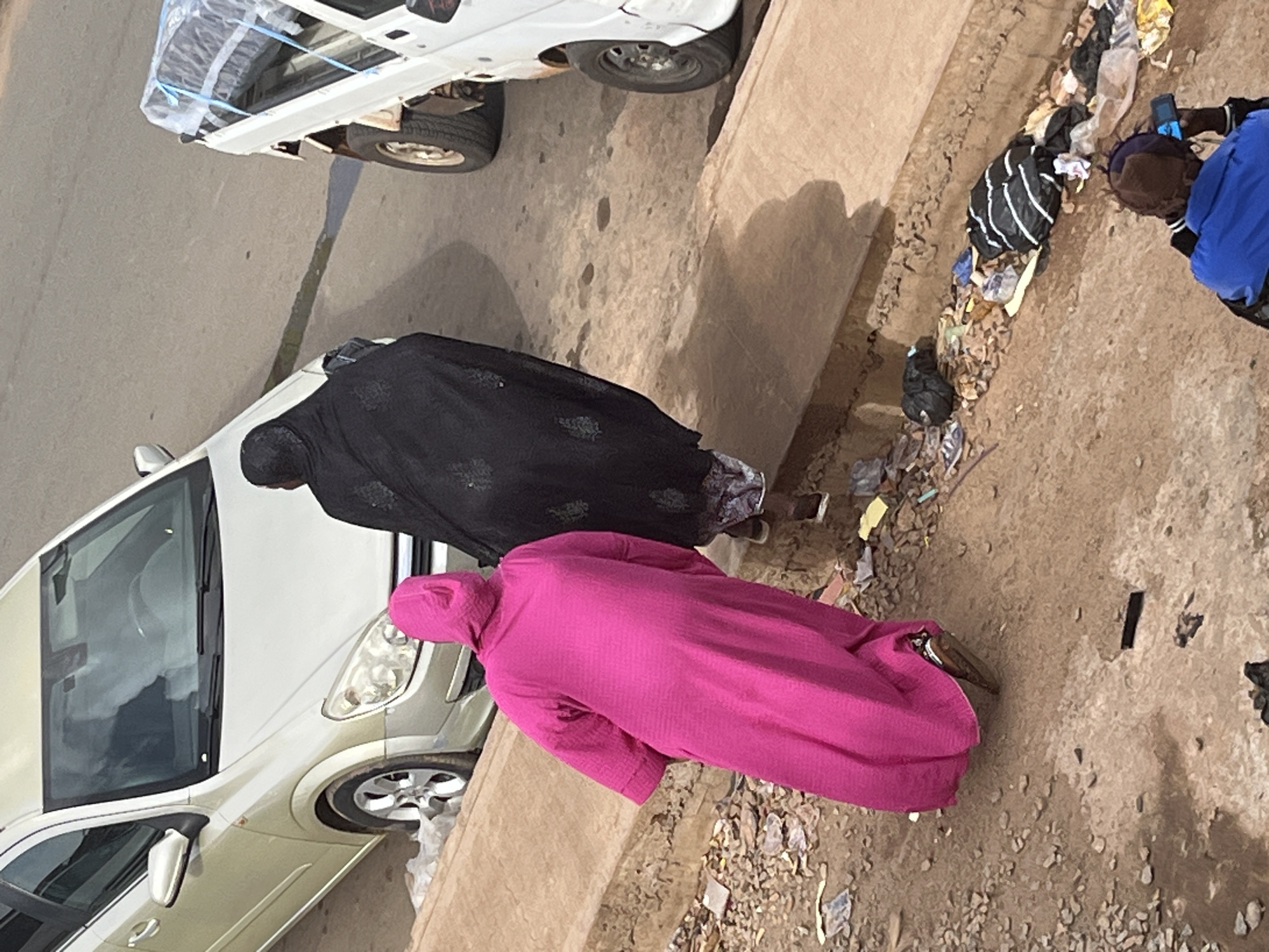 A high-angle, full shot shows two women walking away from the camera on a dirt roadside in Jos, Nigeria. The woman on the left is wearing a bright pink, hooded robe, and the woman on the right is wearing a black abaya. They are walking past a parked silver sedan. To the right of the sedan is a white pickup truck with its bed covered by a tarp. The ground is covered with loose dirt, gravel, and trash, including plastic bags and wrappers. A person is visible in the bottom right corner of the frame, looking at a phone.