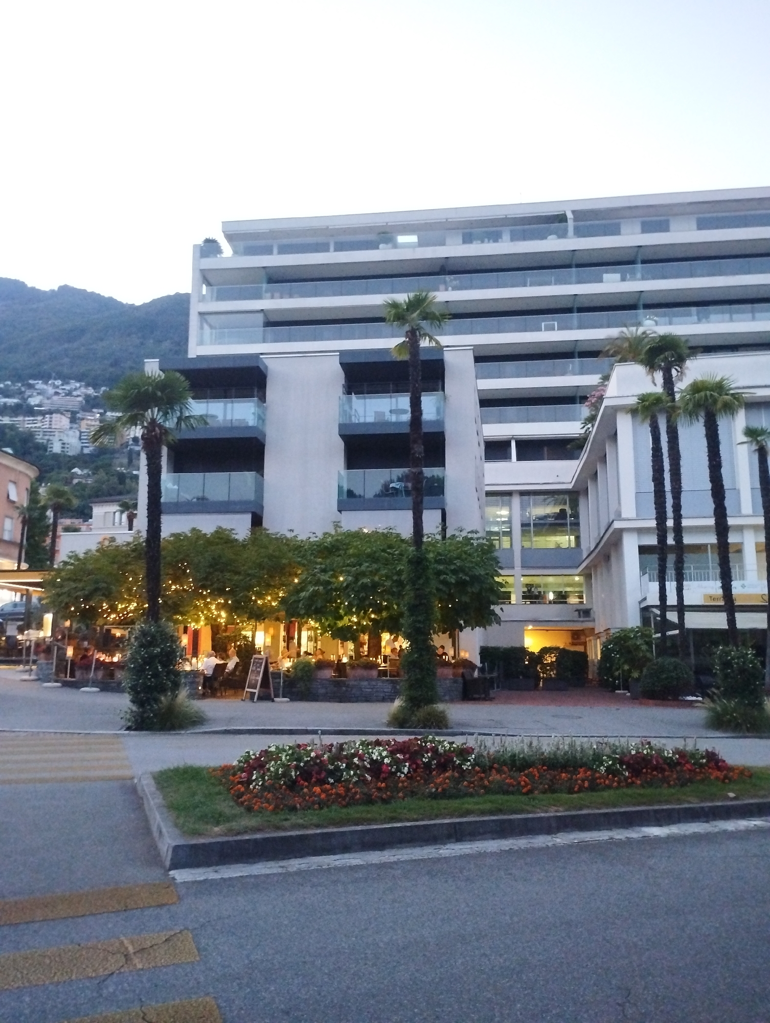 The image captures a street scene in Muralto, Switzerland, featuring a modern, multi-story building with balconies overlooking a street lined with trees and outdoor dining areas. The building's facade is predominantly white with large glass windows and railings. In the foreground, a pedestrian crossing with yellow stripes is visible on the asphalt road. Adjacent to the crossing is a raised flowerbed filled with red, white, and orange flowers. Several palm trees are scattered throughout the scene. An outdoor seating area, illuminated by string lights, is set up in front of the building, with people seated at tables. The background shows a hill with buildings and vegetation. The sky is overcast and the overall lighting suggests it is either early morning or late evening.