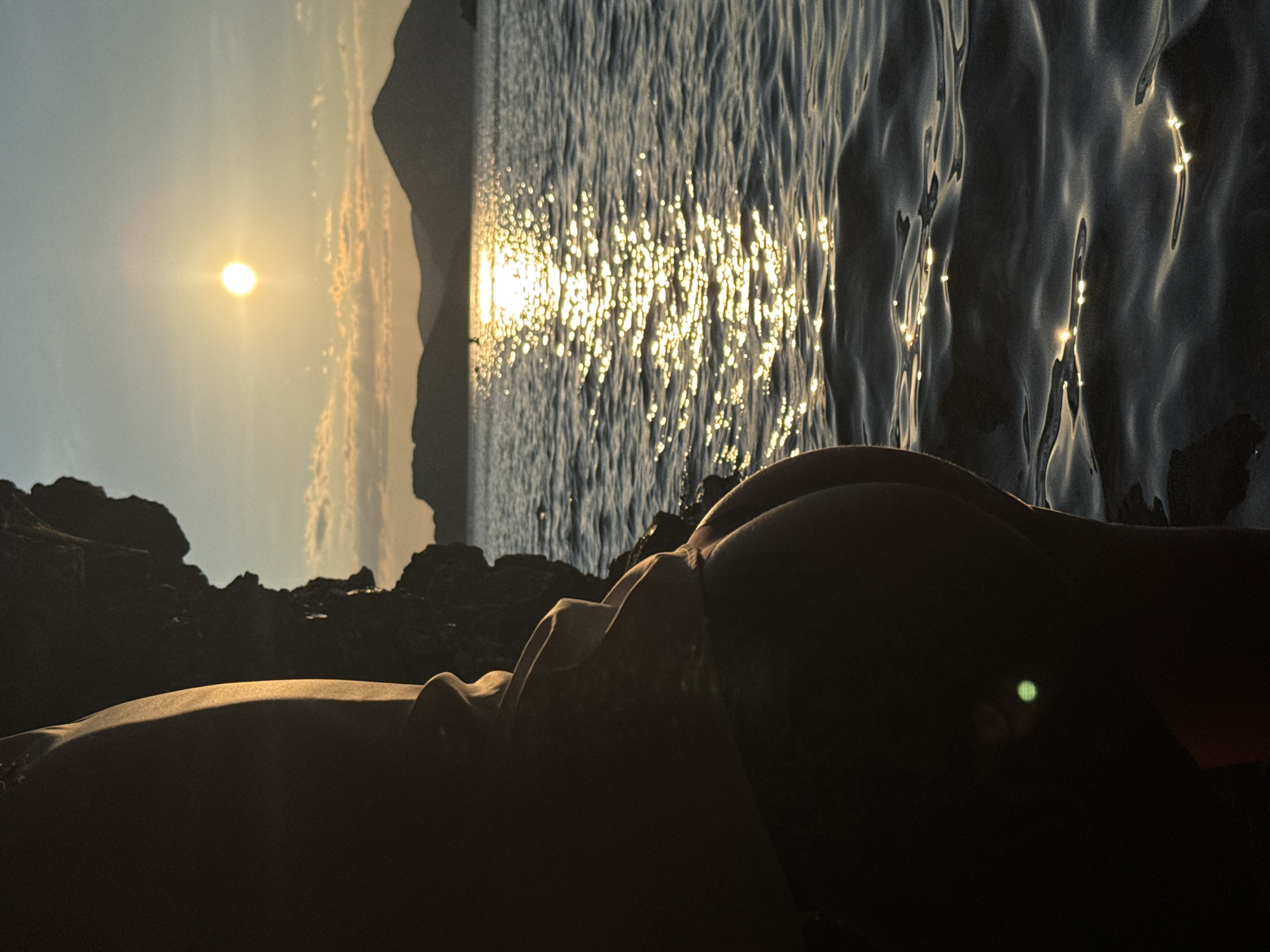 The image shows a person partially visible in the foreground, their back to the camera, positioned next to a dark, rocky formation. The person is silhouetted against a scenic backdrop of a body of water reflecting the golden light of a setting sun. In the distance, rolling hills or mountains are visible against a hazy sky with scattered clouds. The water is choppy, with highlights from the sun creating a shimmering path across its surface. The scene is consistent with a location like Sveti Stefan, Montenegro, known for its beautiful coastline and islands. The overall impression is one of a tranquil evening at the seaside.