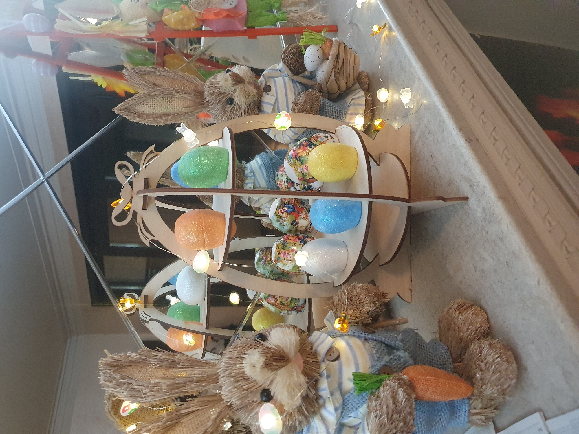 The image shows an indoor Easter decoration, likely in Plovdiv, Bulgaria (inferred due to location prompt). The primary focus is a multi-tiered wooden Easter egg display stand holding various brightly colored Easter eggs. Two straw bunnies flank the egg display. One is dressed in blue and white striped pajamas and carries a carrot. Fairy lights are intertwined throughout the display. The scene is mirrored, suggesting the items are placed on a reflective surface like a glass table. Background elements include parts of a shelf with other decorative items and a glimpse of a fireplace or mantle.