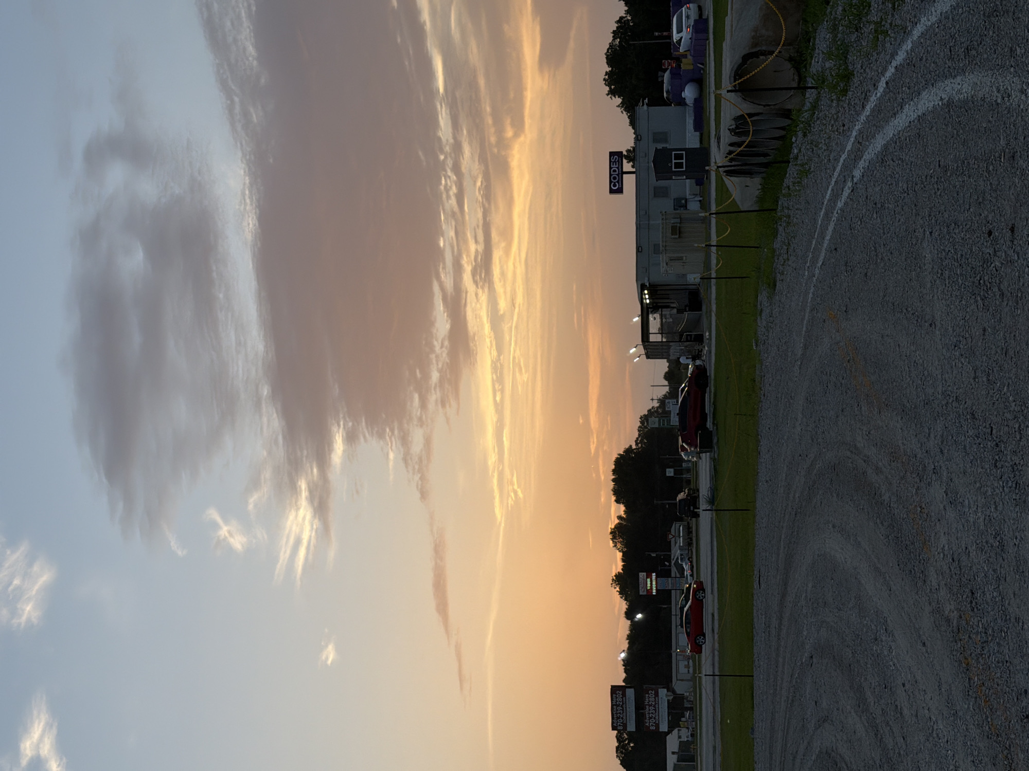 A dusk scene captures a gravel lot in Cardwell, United States, under a sky with wispy clouds tinged with sunset orange and pink. In the foreground, white painted lines delineate a curved path on the gravel. Beyond the lot, a line of trees forms a dark silhouette against the horizon. Several buildings and signs are visible, including a structure with a "CODES" sign and other modular buildings. Cars are parked or driving on a road in the middle ground, and large concrete pipes are stacked along the edge of the gravel area. The overall atmosphere is calm, with the fading light creating a soft, diffused illumination.