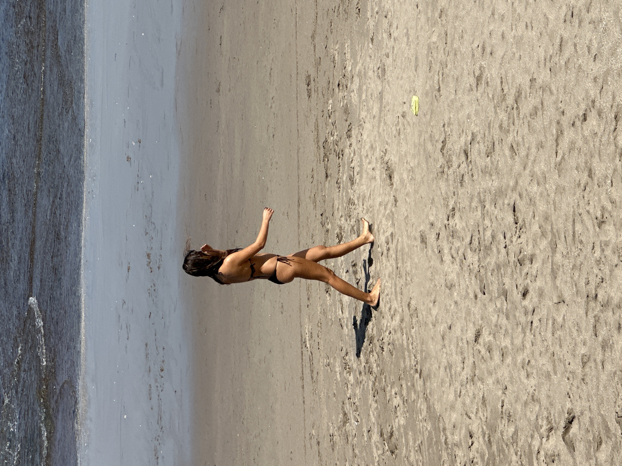 A person is on a sandy beach, near the water. They are wearing a dark bikini and their hair is dark brown. The person's arms are extended, and their body is angled as if they are playing or dancing. The sand has a rough texture, with small indentations and pebbles. To the right of the person, there is a small, bright yellow object on the sand, possibly a toy or piece of litter. The water is a dark grey-blue and appears to be gently lapping at the shore. The lighting suggests it is daytime, likely sunny, as there is a distinct shadow cast by the person. The overall scene is one of leisure and outdoor activity at the beach.