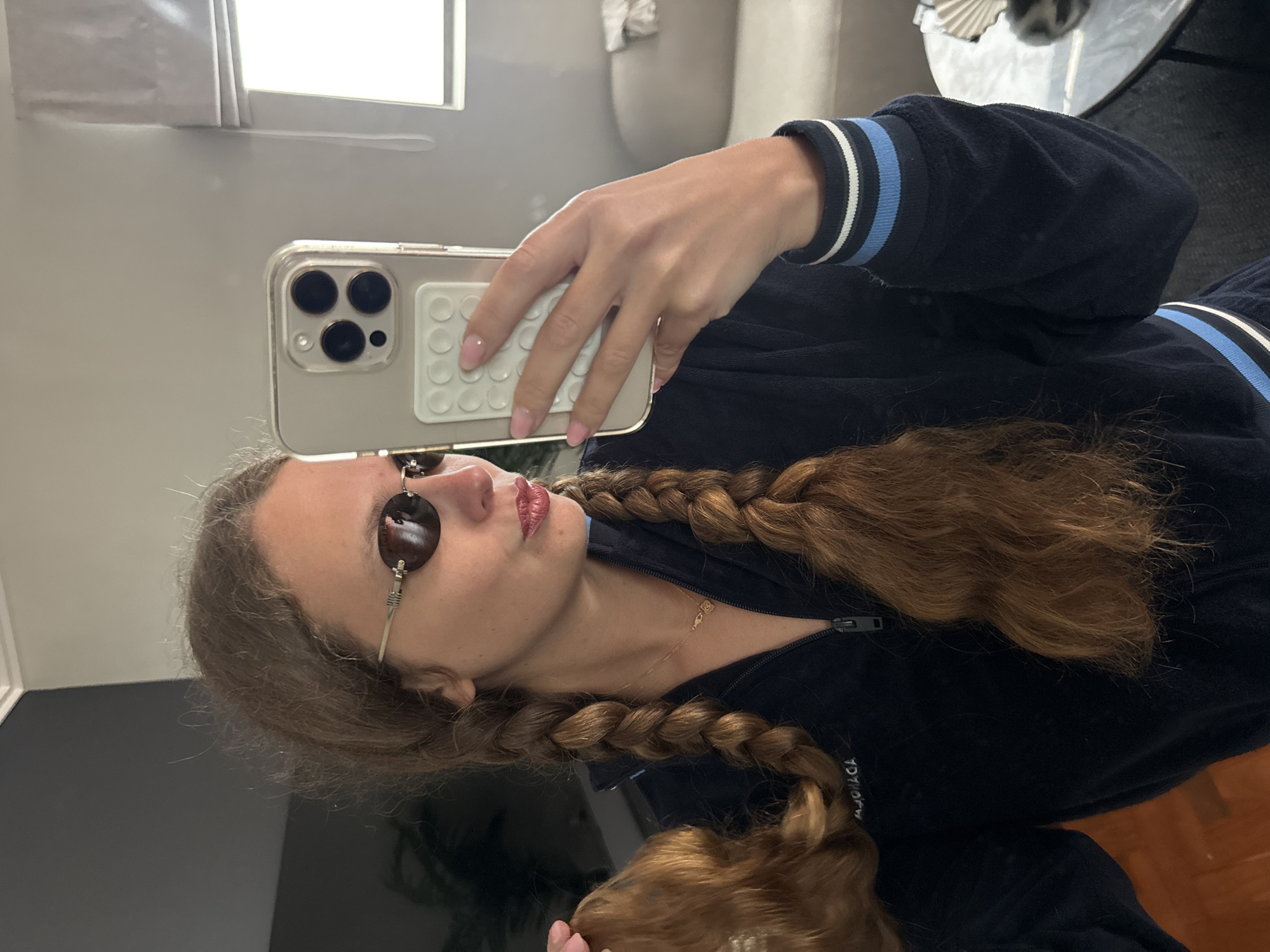 A person with long brown hair styled in two braids is taking a selfie in what appears to be an indoor environment. The person is wearing sunglasses and a dark, possibly navy blue, zip-up jacket with light blue and white striped cuffs. They are holding a smartphone with a clear case and a white adhesive grip on the back. The background includes a window with light-colored curtains, a sofa, and a round table with a dark surface. The image does not contain sufficient information to definitively determine if the location is in Cape Town, South Africa.