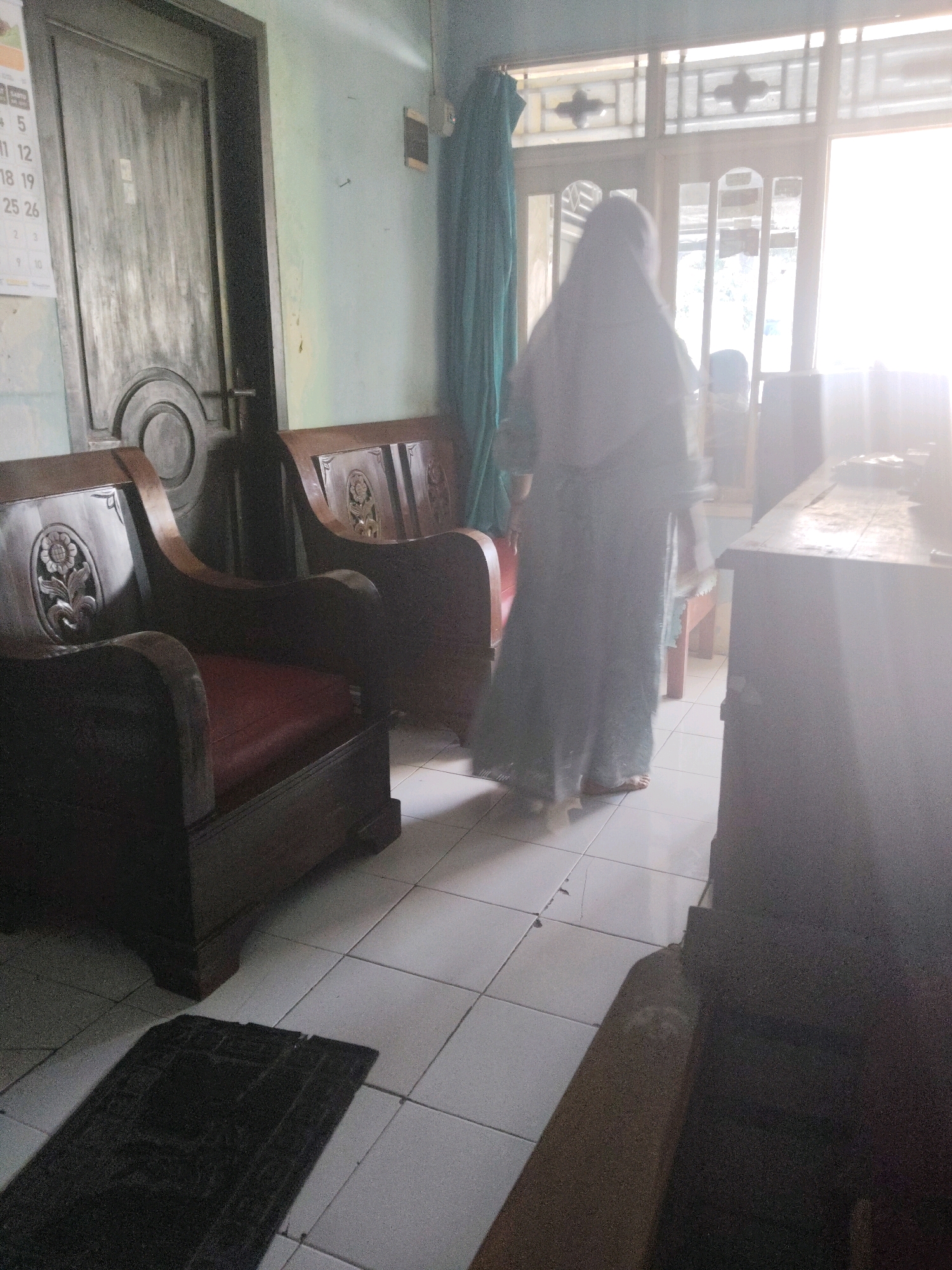 The image depicts an interior scene, likely a living room, located in Subang, Indonesia. A person wearing a hijab and a long dress stands facing away from the camera, towards a window with natural light streaming in. The room features traditional wooden furniture, including a carved sofa and another chair. A doorway with a calendar on the left side of the frame indicates the presence of an entrance. The floor is tiled. To the right of the frame, a large, dark cabinet is visible, partially obscuring the view of the cabinet's surface. A small prayer mat is placed on the floor in the foreground.