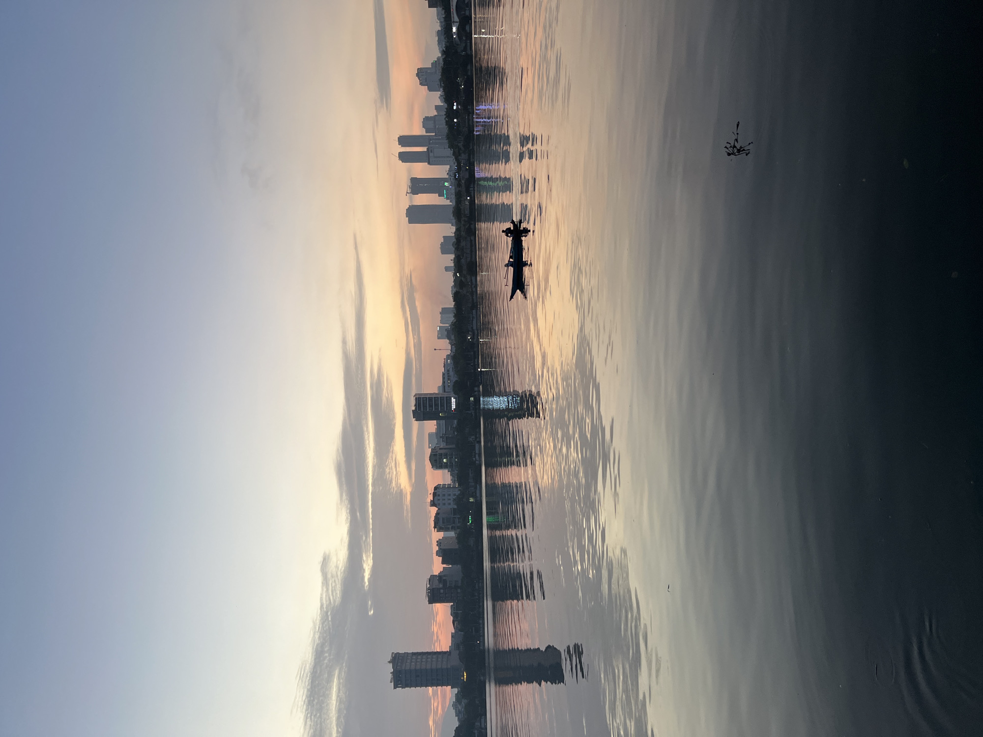 This is a serene, low-angle shot of a lake with a cityscape in the background, likely taken at dawn or dusk. The sky transitions from a pale blue at the top to hues of orange and yellow near the horizon. Wispy clouds are scattered across the sky.
The water's surface is calm, with a clear reflection of the buildings and the sky. A small, dark boat with two figures silhouetted inside is positioned in the middle of the lake. The boat appears to be moving slowly.
In the distance, a skyline of various buildings, including some tall, modern structures and smaller, older ones, is visible. The architecture suggests an urban environment. A reflection of the city lights and buildings shimmers on the water.
The lower portion of the image is dominated by the dark, still water, with subtle ripples. A small, dark object, possibly a plant or debris, is floating on the water's surface towards the bottom right.
While the image shows an urban setting, the presence of a lake and the specific skyline architecture would be needed to definitively pinpoint the location in Da Nang, Vietnam. However, the scene evokes a tranquil atmosphere characteristic of waterside urban areas, potentially such as Han River in Da Nang.