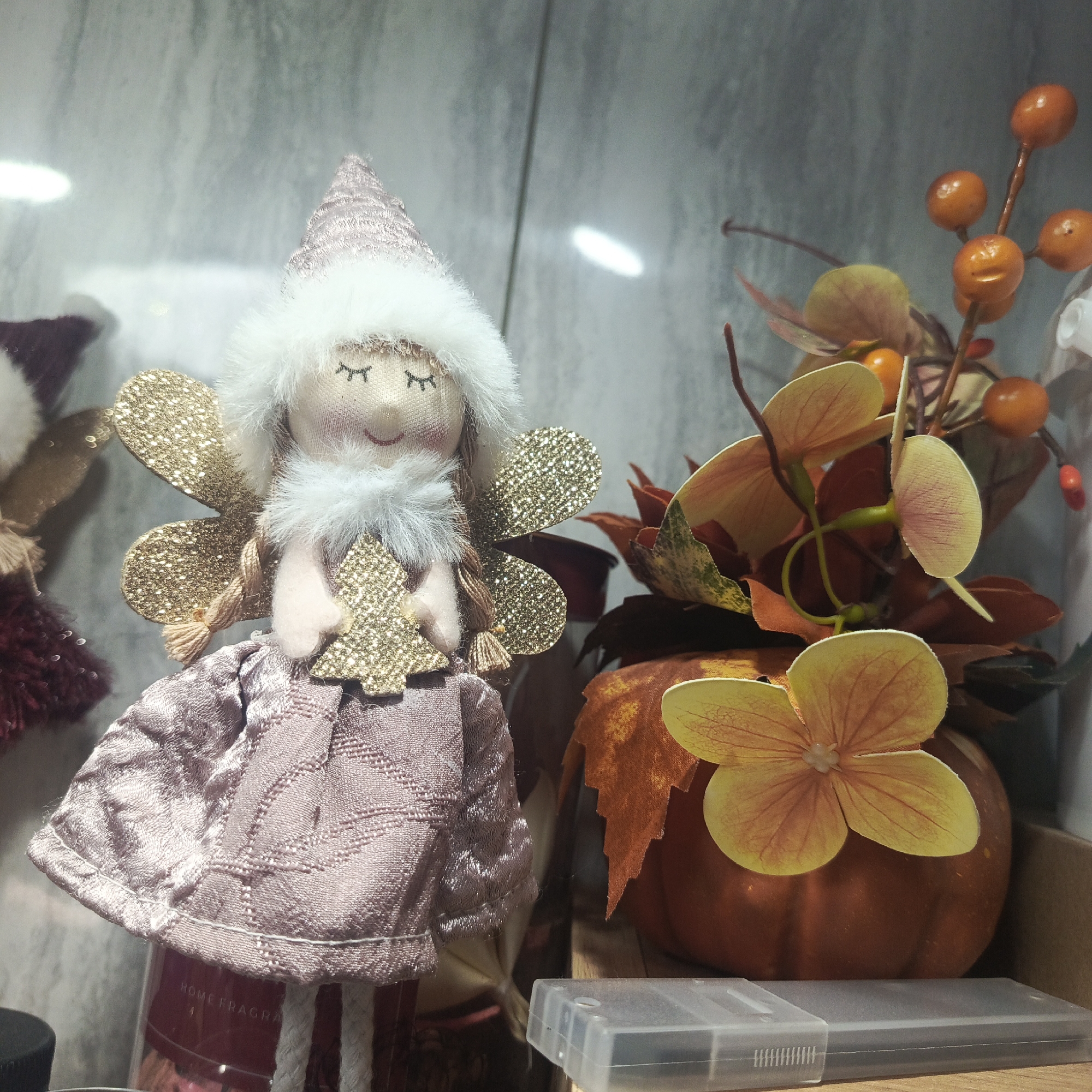 A fairy doll and a decorative pumpkin with autumnal floral decorations are situated within a confined, likely enclosed, space. The doll has a pink fabric body, white fur trim, glittery gold wings, and closed eyes. The pumpkin is orange and embellished with foliage and orange berries. A translucent plastic container sits at the base of the pumpkin. The location, Марковский, Russia, is contextually extraneous and not directly discernible from the image.