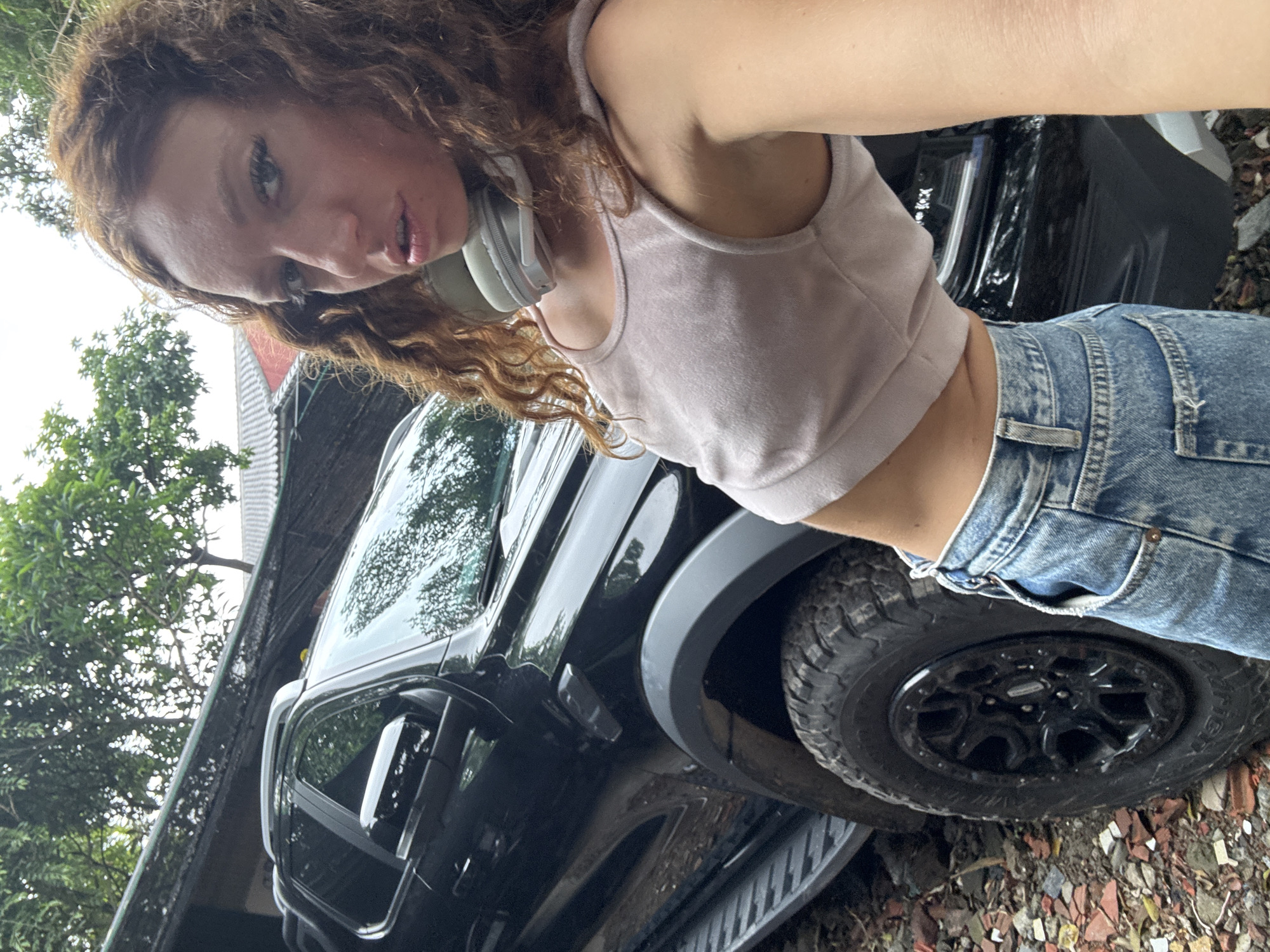A red-haired woman stands next to a black pickup truck in Hà Nội, Vietnam. She wears a light pink tank top, high-waisted blue jeans, and white headphones around her neck. The truck is parked on a partially paved surface with scattered debris. Trees and a building with a corrugated metal roof are visible in the background.