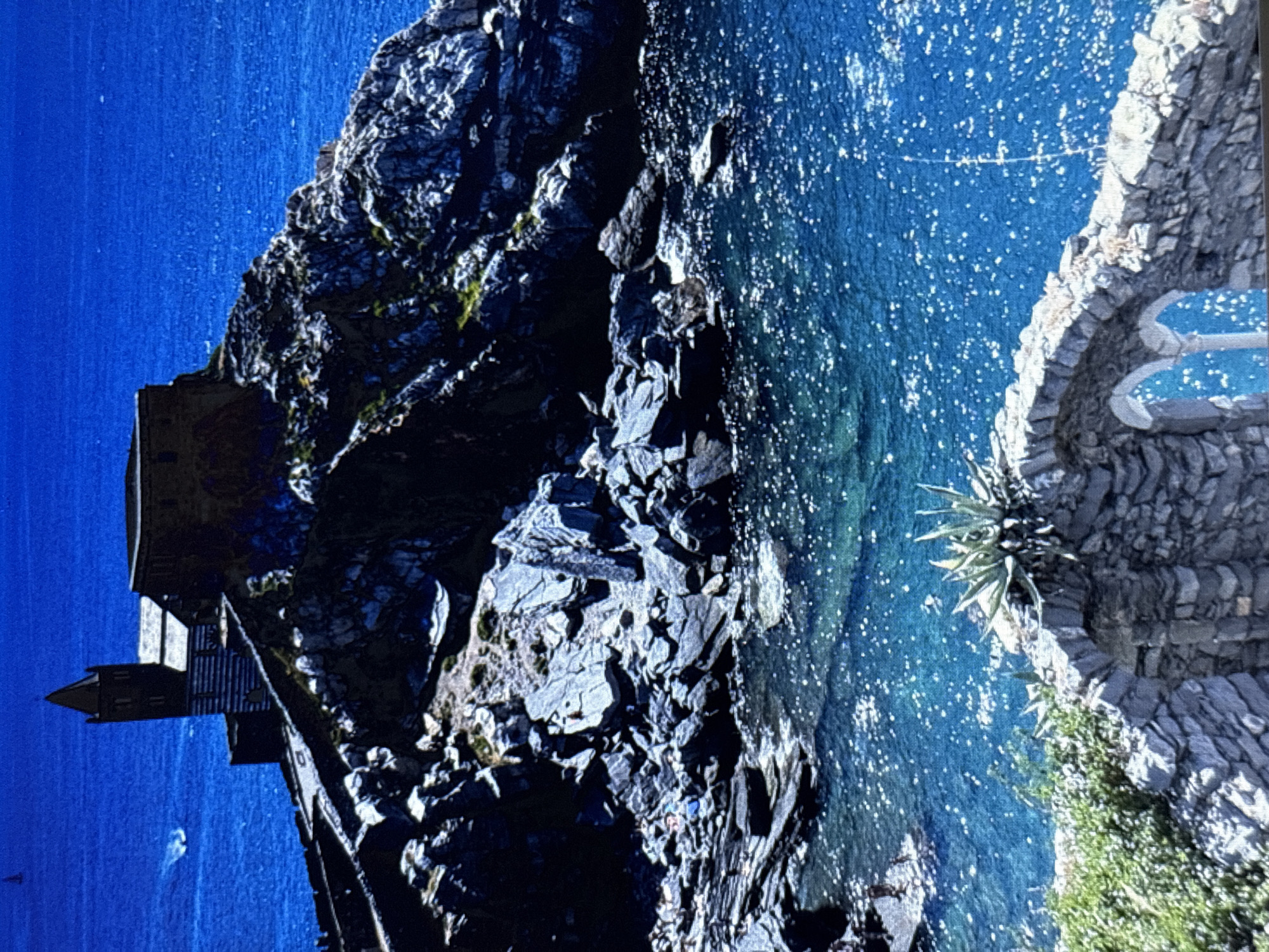 The image shows a striking cliffside scene with a building perched precariously on the edge. The building appears to be made of stone and has a dark, imposing facade. It is situated above a turbulent, deep blue body of water, possibly the sea or a large lake. The water is choppy, with whitecaps indicating some wind or swell. Scattered across the water's surface are numerous bright white specks, possibly reflections of sunlight or spray.
The cliff face is rugged and irregular, composed of dark gray rocks with patches of green vegetation clinging to its surface. The texture of the rocks suggests a natural, weathered formation. On the right side of the image, a stone-built terrace or pathway curves along the edge of the cliff. This structure is made of rough-hewn stones and leads to two distinct, blue-colored shapes that could be decorative pools or seating areas. A spiky, desert-like plant grows from a crevice in the stone wall.
The overall impression is one of dramatic natural beauty and human resilience, with the building clinging to the edge of a formidable landscape. The time of day appears to be daytime, given the bright sunlight reflecting off the water and highlighting the textures of the rocks and the building. The weather seems to be windy, as suggested by the state of the water. The location is identified as Sesto Calende, Italy. There is no visible text in the image.