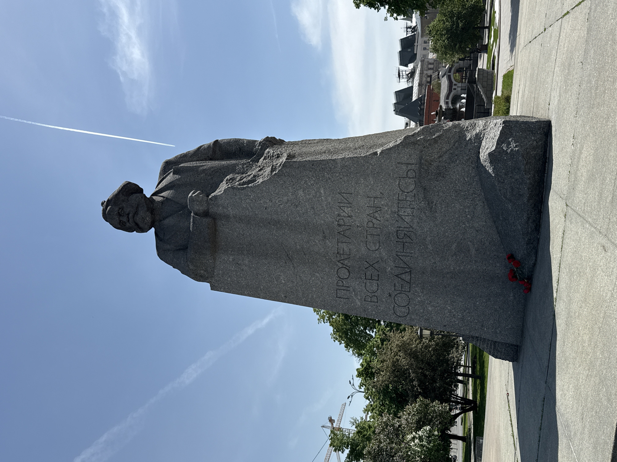 The image shows a granite statue of Karl Marx in Moscow, Russia. The inscription on the statue reads "Proletarians of all countries, unite!" in Russian. Red flowers are placed at the base of the statue. In the background, trees and buildings can be seen under a blue sky. A contrail cuts across the sky.