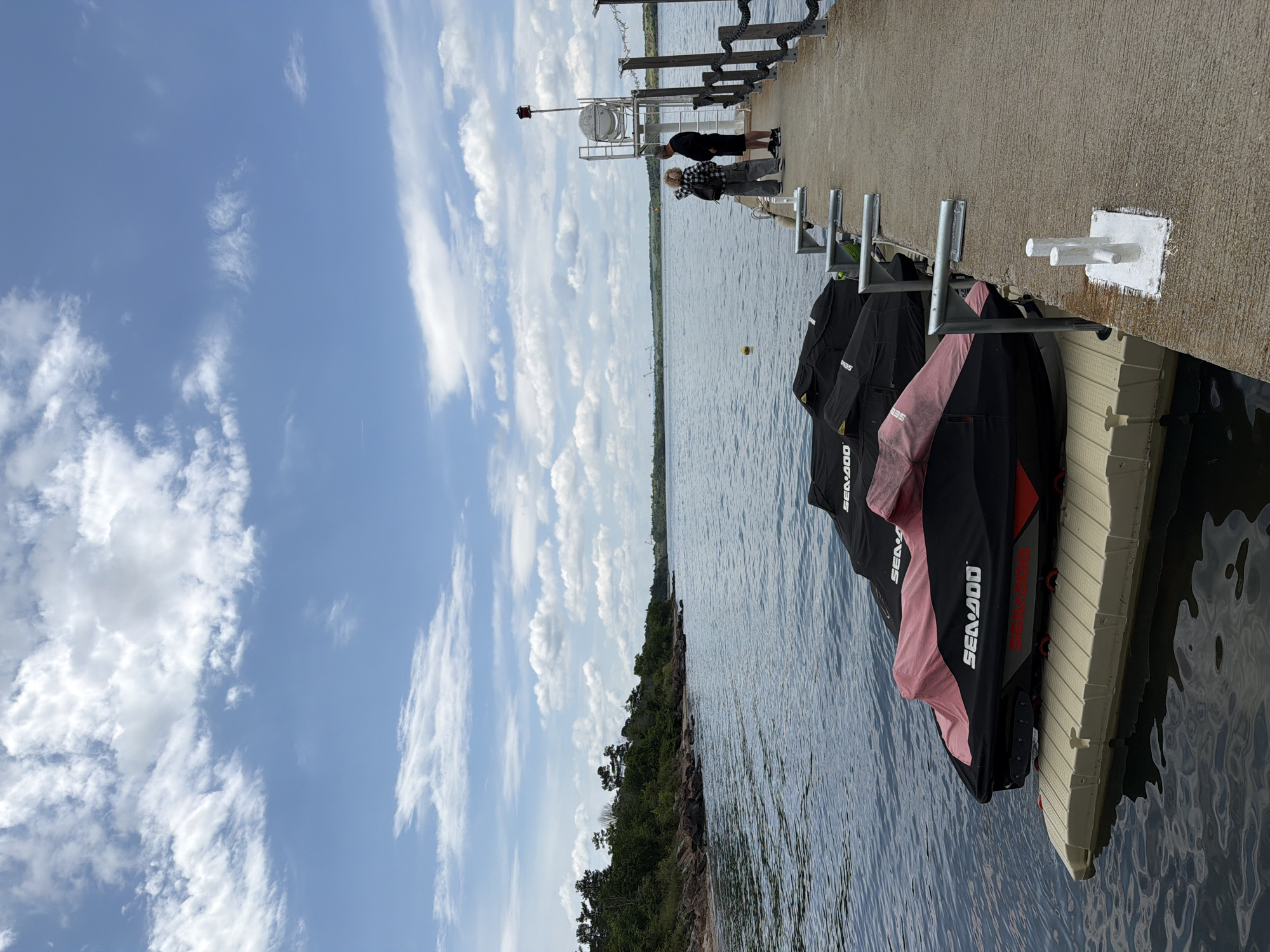 The image shows a waterfront scene with several personal watercraft (jet skis) docked along a concrete pier. The jet skis are black with some having pink covers. The pier has metal and white-painted mooring posts. Two people are standing on the pier towards the center of the frame. The sky is blue with scattered white clouds, and there is a body of water with a tree-lined shore in the background. The scene appears to be a sunny day at a recreational area. The location is not identifiable as Burgas, Bulgaria from the image alone.