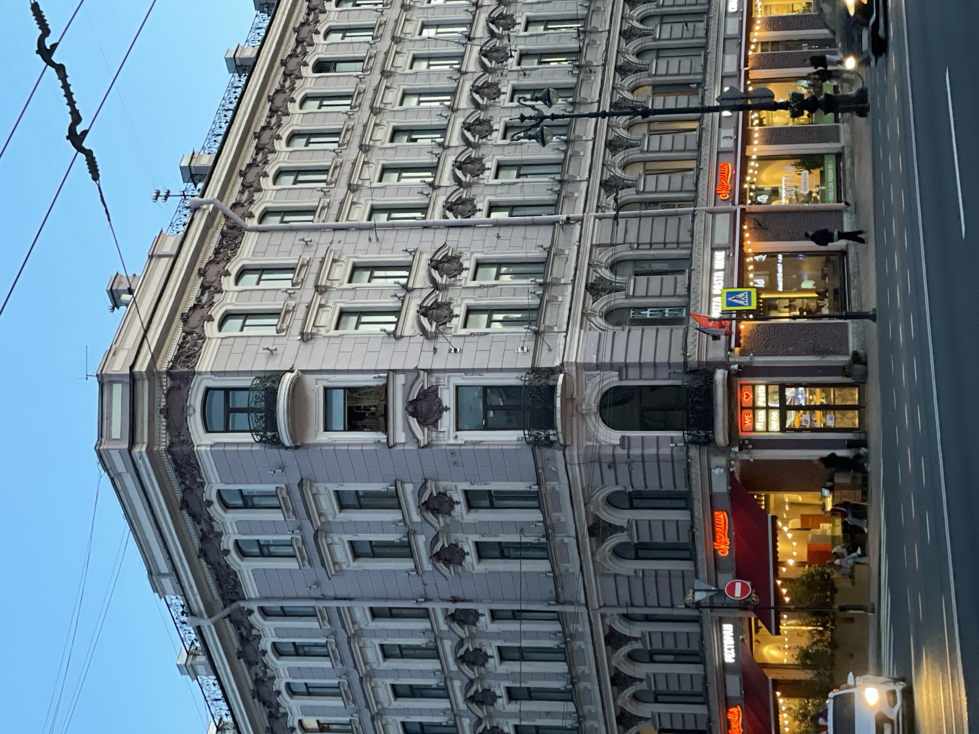 The image captures a detailed view of a multi-story ornate building facade along a city street in Saint Petersburg, Russia, at dusk or early evening. The sky, a clear light blue, appears above the building, crisscrossed by overhead power lines.
The building is constructed of light-colored, possibly grey or beige, stone or stucco, showcasing rich architectural details. It features numerous rows of windows, many with arched tops or elaborate decorative surrounds and balustrades between floors. Several balconies with intricate wrought-iron railings extend from the upper levels. Ornate cornices and decorative elements are consistently present across the facade, indicating a historical and elaborate architectural style.
The ground floor houses several commercial establishments, likely restaurants or cafes, distinguished by large, brightly lit windows and prominent red awnings. Visible text includes "Маргарита" (Margarita), appearing multiple times, along with "РЕСТОРАН" (Restaurant). Other signs, partially visible, read "PIZZA PASTA WINE" and "WE LOVE PIZZA" in English. The warm interior lighting suggests these businesses are open and active.
A wide sidewalk runs in front of the establishments. One person is clearly visible walking on the sidewalk. Adjacent to the sidewalk is a paved street with a white lane marking. A white vehicle with illuminated headlights is partially visible on the street, suggesting ongoing traffic. Ornate streetlights with multiple lamps illuminate the street and sidewalk. Traffic signs, including a blue pedestrian crossing sign and a red circular "no entry" sign, are also visible. The overall scene depicts a vibrant urban environment as day transitions into night.