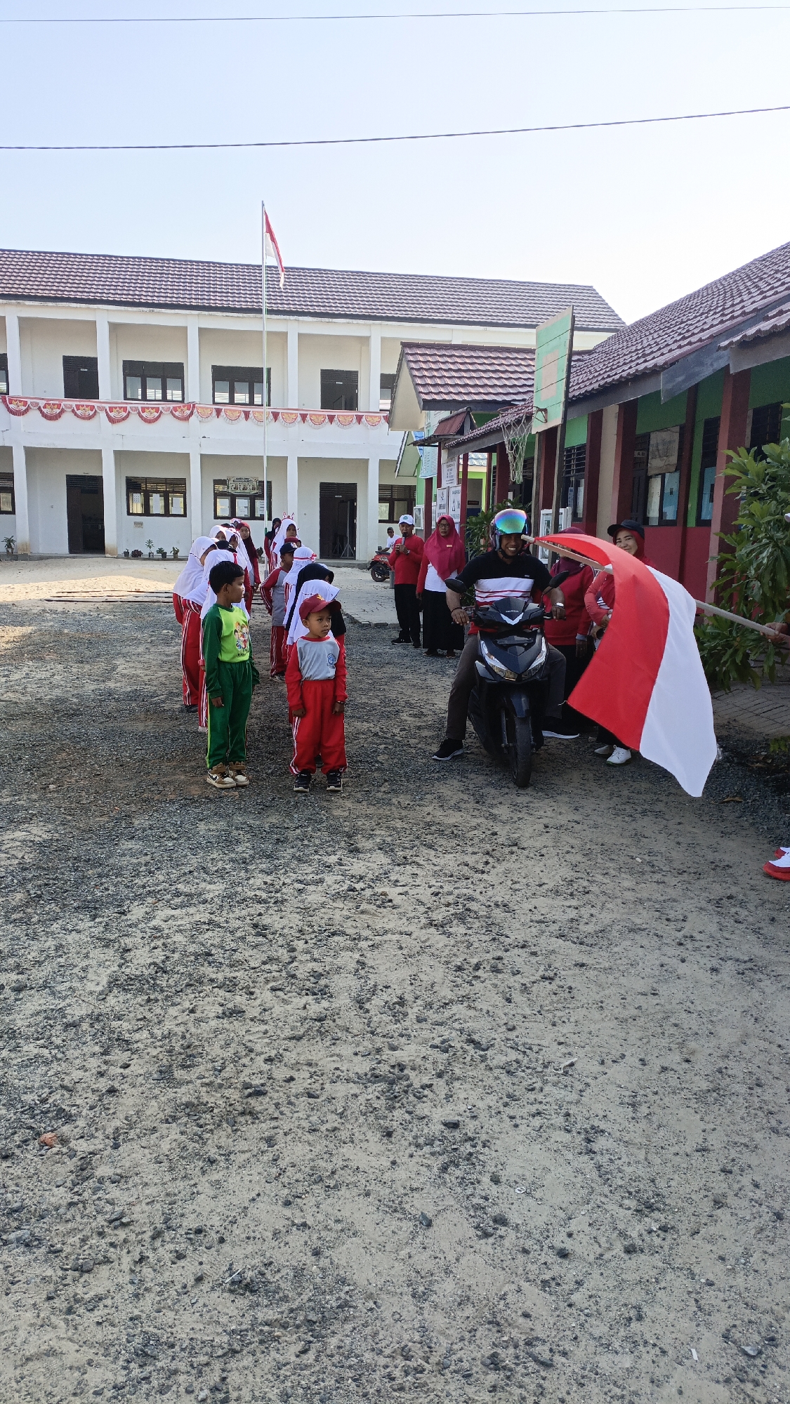The image depicts an outdoor scene in Saring Sungai Binjai, Indonesia, possibly at a school. In the foreground, two young boys are standing, one in a green uniform and the other in a red and grey uniform. Behind them, a group of children in red and white uniforms are lined up. To the right, a man wearing a helmet is riding a black motorcycle. He is holding a large Indonesian flag, which is red and white, extending to the right side of the frame. Several adults, some in red clothing, are standing near the motorcycle and the flag. The background shows a long building with a tiled roof, a flagpole with a red and white flag, and what appears to be another building with green trim. The ground is covered in gravel. The sun is shining, casting shadows.