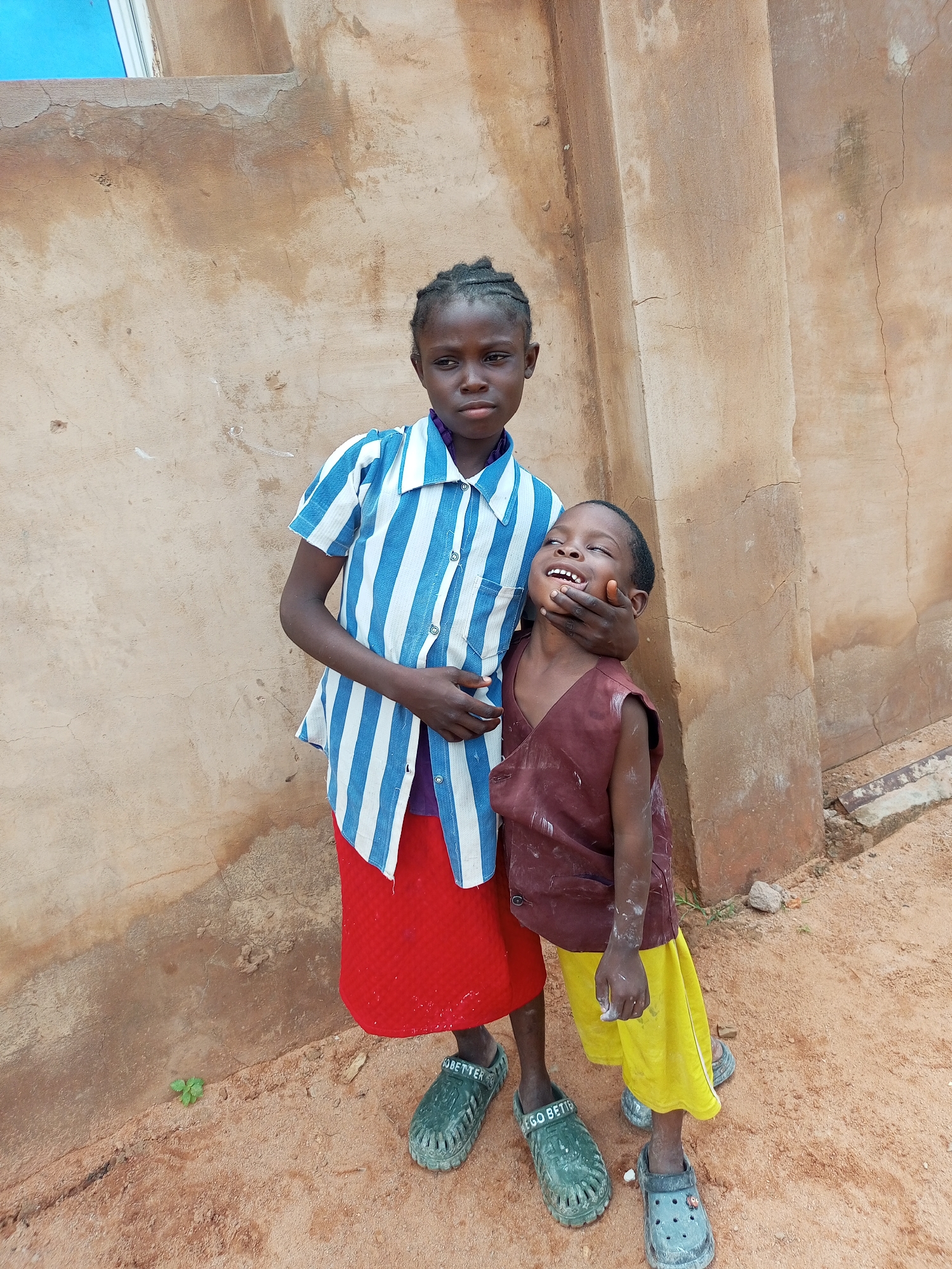 Two children, a girl and a boy, stand against a textured, light brown wall in what appears to be Tempure, Nigeria. The girl, standing on the left, is wearing a blue and white striped button-down shirt over a purple undershirt and a red skirt. Her hair is styled in braids. The boy, on the right, is wearing a sleeveless maroon tunic over yellow shorts. His head is tilted back and his mouth is open, appearing to laugh or call out, with the girl's hand resting on his jaw. Both children are wearing casual shoes. The ground beneath them is composed of reddish-brown dirt and sand, with a few small green plants growing in the dirt. The wall behind them shows signs of wear and cracking.