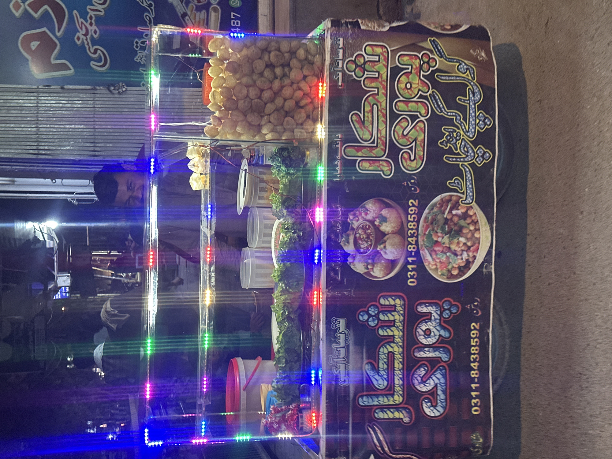 Here is a detailed description of the image:
**General Description:**
The image shows a food cart, likely a street vendor stall, illuminated with vibrant LED lights. It appears to be taken at night. The overall impression is of a lively food service operation.
**Objects and Details:**
* **Food Cart:** The cart has a multi-tiered display case, filled with various ingredients and food items.
* Top tier contains fried balls or spheres, possibly chickpea balls.
* Lower tiers hold containers of condiments, salads, and ingredients.
* **LED Lights:** Multicolored LED strip lights illuminate the perimeter of the display case, adding a festive atmosphere. Colors include blue, purple, green, yellow, and red.
* **Signage:** The cart has signs in what appears to be Urdu script. Phone numbers (0311-8438592) are prominently displayed, indicating this is a business advertisement.
* **Food items visible:** The cart contains fried puffed puri, as well as Chana Chat.
**People:**
* **Vendor:** The reflection of a person, likely the vendor or operator of the cart, is visible in the glass of the display case. They appear to be wearing glasses and a dark jacket or shirt.
**Environment:**
* **Location:** The setting appears to be an urban environment. This is suggested by the presence of other businesses in the reflection.
* **Time:** The image was taken at night.
**Overall Impression:**
The image captures a vibrant street food vending scene. The combination of food, lights, and signage creates an appealing and inviting atmosphere, typical of urban street food culture.