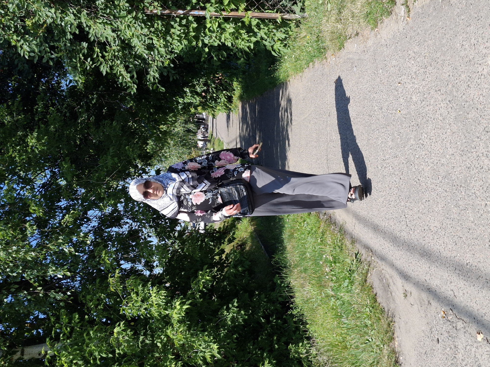 A person wearing a hijab, sunglasses, a floral-patterned long-sleeved shirt, and gray pants walks along a paved path lined with trees and grass. The person is carrying a black crossbody bag and holding a phone in their right hand. The sun is shining, casting a shadow on the path. The path appears to be in a residential or park area.