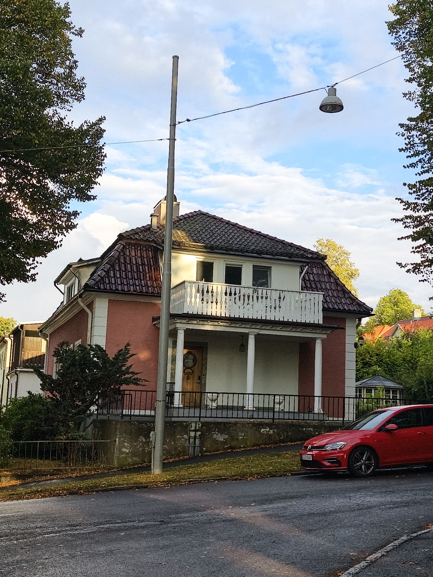 This is a photograph taken in Norrköping, Sweden, capturing a suburban street scene with a distinctive house and a red car. The house, a multi-story structure with a salmon-pink facade and a dark tiled roof, features a prominent white balcony with ornate railings supported by columns. A wooden door is visible on the ground floor. The house is bordered by a black metal fence and a low concrete wall. Adjacent to the house, a dark green evergreen shrub grows. A tall, slender utility pole stands in front of the house, with a wire extending from it to a streetlamp hanging over the road. The road itself is asphalt with visible wear and tear, lined with fallen leaves. A red Volkswagen Golf is parked on the right side of the road, facing away from the camera. The background includes lush green trees and a partially visible gazebo. The sky above is a mix of blue with scattered white clouds, suggesting an overcast day.