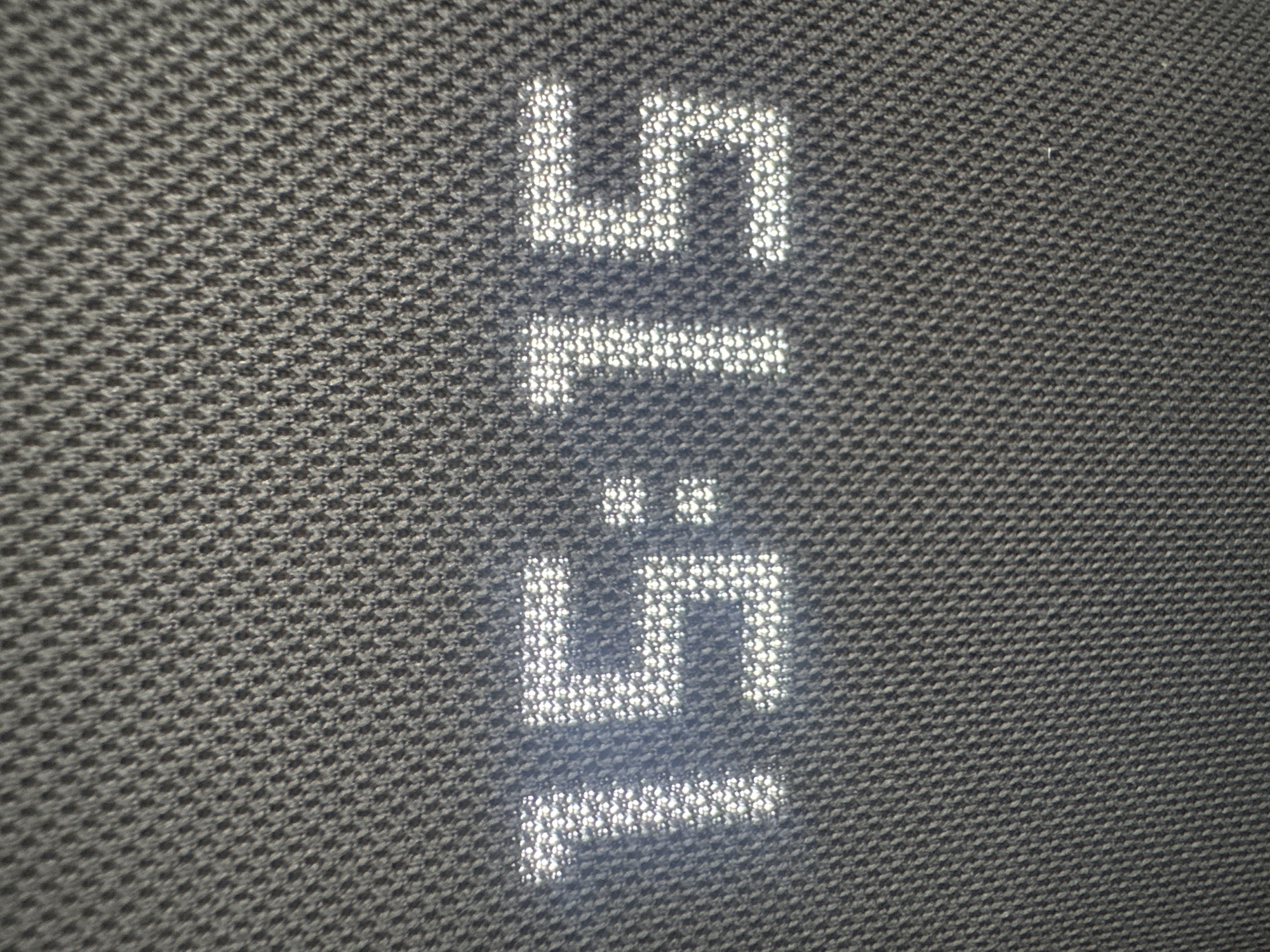A close-up shot captures an illuminated digital display on a dark, textured fabric surface. The fabric exhibits a fine, repeating woven pattern, possibly a houndstooth or similar small-scale weave, giving it a mottled grey appearance.
The primary subject is the bright white text, which appears to be "15:15" displayed twice in a vertical arrangement. Each digit and the colon are formed by numerous small, illuminated dots or pixels, suggesting an LED display embedded within or shining through the fabric, or reflected upon it. The characters are blocky and distinct, contrasting sharply with the dark background. The upper set of "15:15" is positioned above the lower set of "15:15".
There are no people or other discernible objects present. The image provides a detailed view of the interaction between the digital display and the textile surface. This setup might be part of a smart speaker, clock, or another electronic device with a fabric covering. The specific location is Krasnodar, Russia.