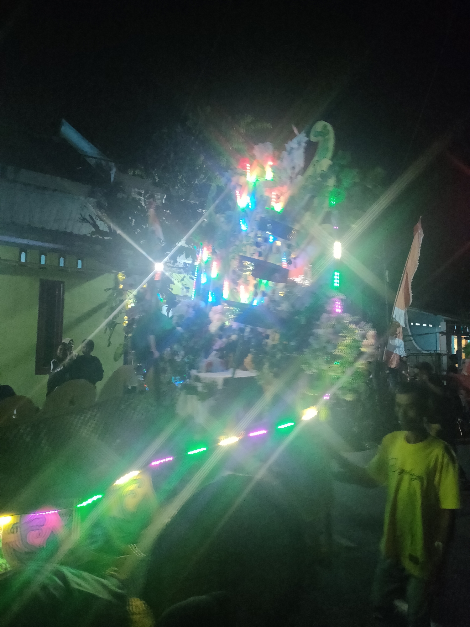 A nighttime scene captures a street celebration in Tongaswetan, Indonesia. The image is dominated by a large, elaborately decorated float or structure adorned with numerous colorful lights. These lights, in shades of green, pink, blue, and white, create a vibrant and festive atmosphere, with some emitting lens flares that streak across the frame. In the foreground, a young man wearing a yellow t-shirt and dark pants stands facing right, partially obscured by the light. To the left, a building with a pale green facade and a dark-colored door is visible. Several people are gathered in the background, some seated on what appear to be chairs. A flag, predominantly orange and white, is positioned to the right of the float. The ground is dark, indicating it's nighttime. The overall impression is one of a lively local event.