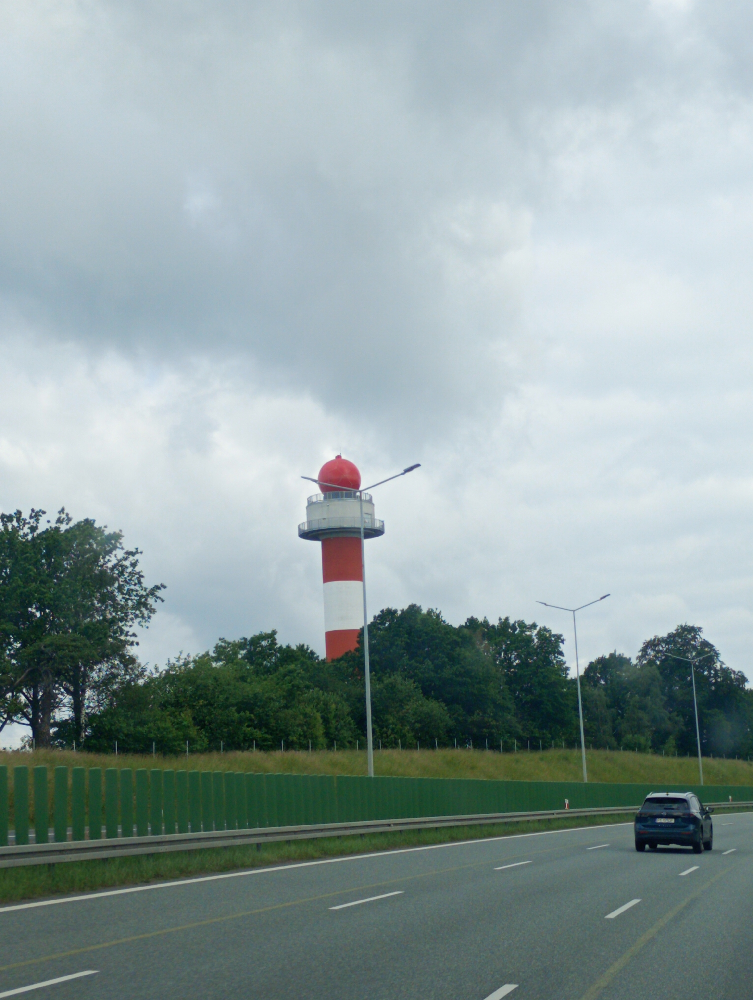 The image captures a view from a highway in Góra Świętej Anny, Poland, looking towards a distinctive red and white tower with a red spherical top, resembling a lighthouse or a similar navigational aid. The tower is situated behind a dense line of green trees. In the foreground, a dark-colored car is driving on the highway, which is marked with white lane lines and a yellow broken line. The sky is overcast with gray clouds. A green noise barrier runs along the side of the highway, separating it from the grassy embankment and trees behind. Several streetlights are visible along the highway.