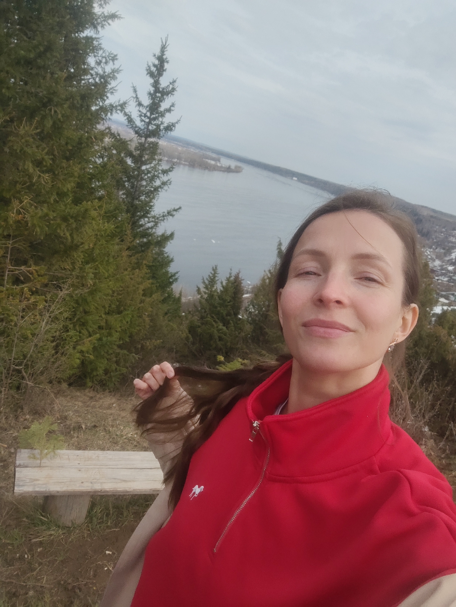 A woman with long brown hair, wearing a red quarter-zip pullover, is taking a selfie outdoors. She's holding a strand of her hair. Behind her is a wooden bench and a variety of evergreen trees. In the distance, a body of water is visible with a town on the opposite shore. Given the location provided (Ольховка, Russia), this body of water is likely the Иловлинское водохранилище (Ilovlya Reservoir). The sky is overcast.