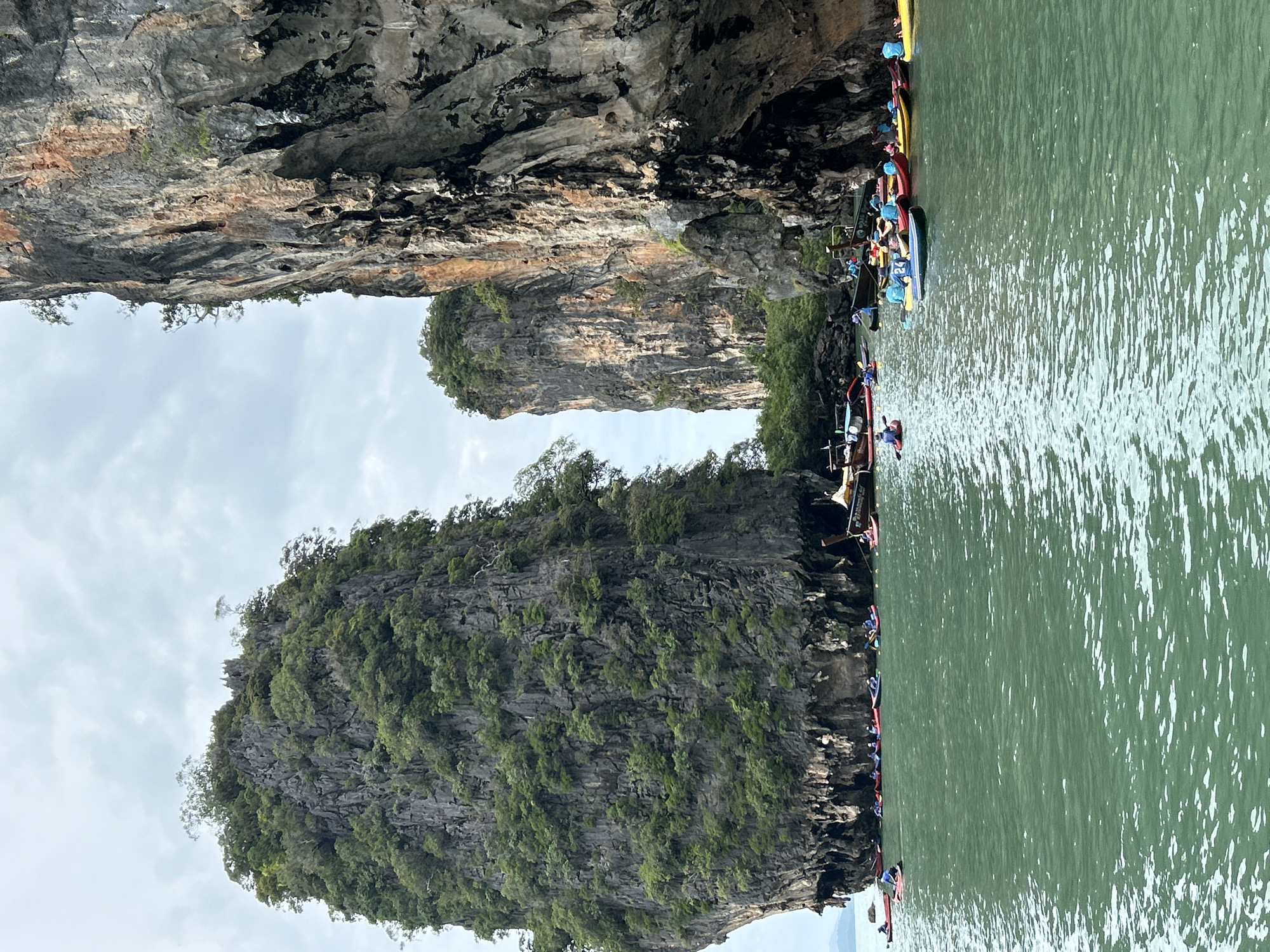 In Krasom, Thailand, a group of people are kayaking in a body of water between towering limestone cliffs. The cliffs are covered in lush green vegetation and are a dominant feature of the landscape. The water is a vibrant green, and several longtail boats are also present, some with passengers visible. The sky is overcast, suggesting a cloudy day. The kayakers are spread out across the water, some in pairs and others individually, appearing to be on a guided tour or recreational outing. The scene depicts a popular tourist destination known for its karst topography and aquatic activities.