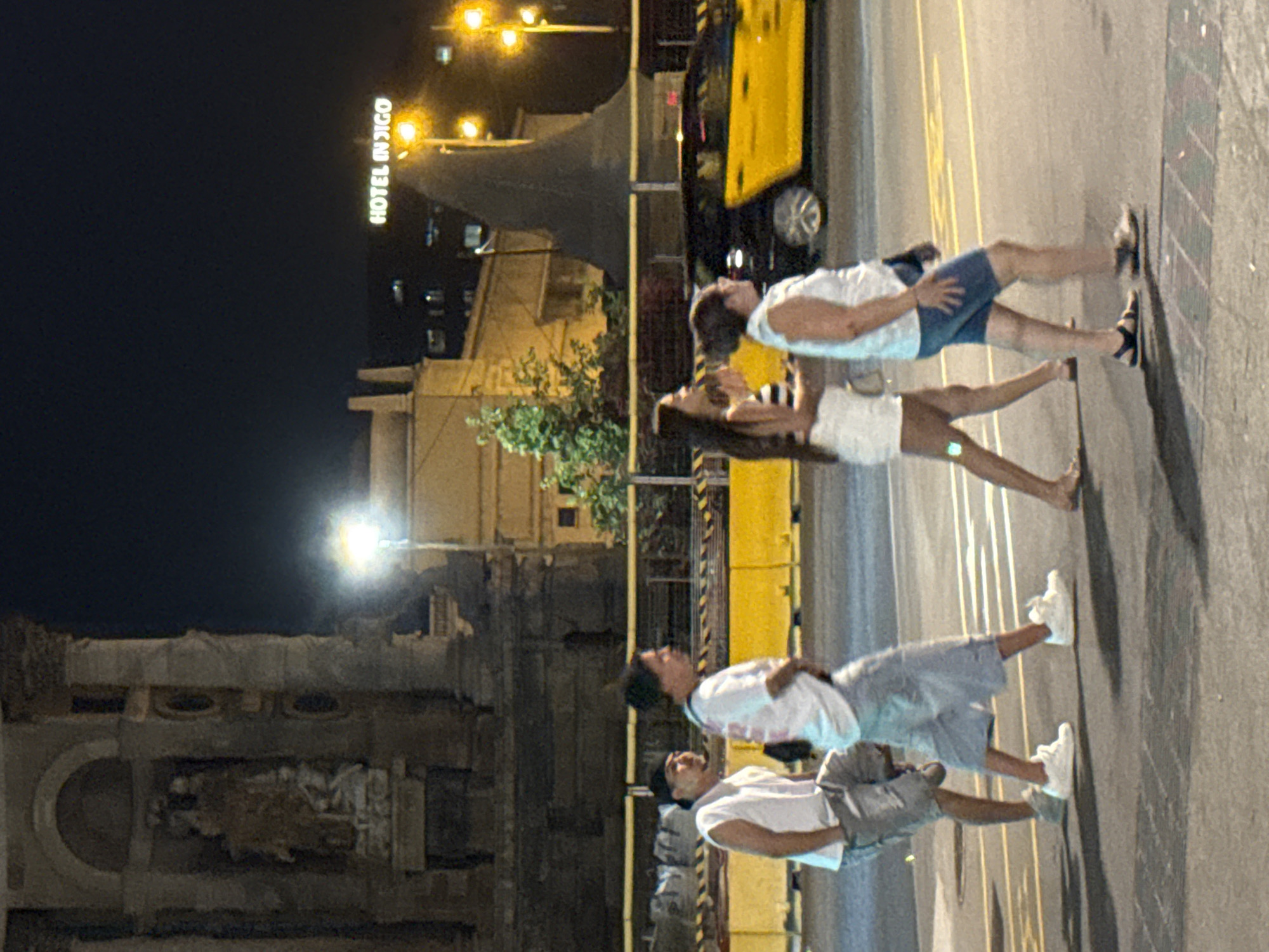 A night-time street scene in Barcelona, Spain, captures four individuals walking from left to right across a crosswalk. The first person, a male wearing a white sleeveless shirt and gray shorts, is looking to the left. Next to him, a male in a white t-shirt and light blue shorts is walking forward. Following them, a female with long dark hair and a striped top is walking, and next to her, a female in a white tank top and blue shorts is also walking. In the background, a yellow taxi is visible, and a building with illuminated "HOTEL INDIGO" signage is present. The environment includes a streetlight and architectural elements of a building on the left side of the frame.