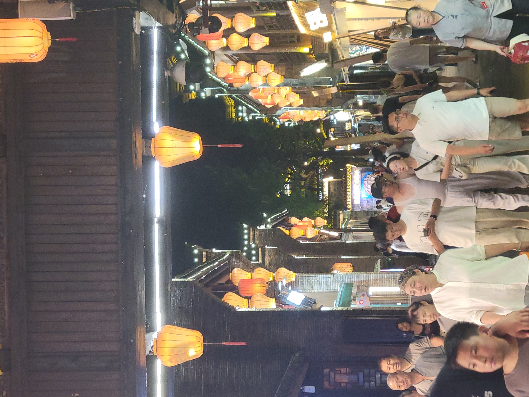The image depicts a nighttime scene on a street in Wuhou District, China. The street is lined with traditional Chinese architecture, illuminated by numerous orange lanterns. A crowd of people are walking along the street, some looking at shops, others engaged in conversation. In the foreground, a young boy is looking to his left, while an adult woman in a white shirt and a patterned headscarf walks forward. Behind them, a man in a white polo shirt and khaki shorts is looking at his phone. Other individuals are visible further down the street, creating a lively atmosphere. Security cameras are mounted on some of the buildings. The overall impression is of a bustling urban environment at night.
