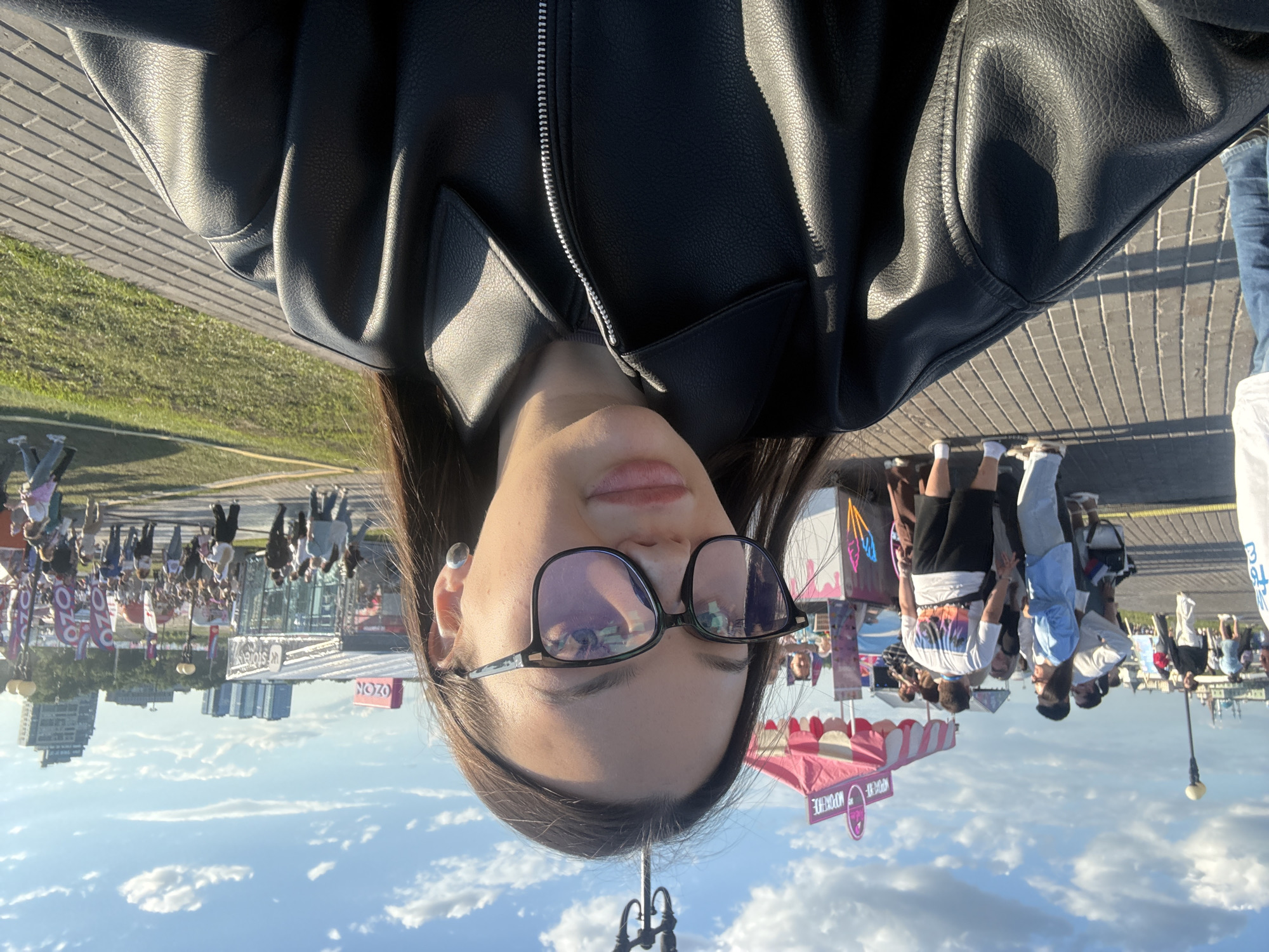 A young woman with long dark hair, wearing a black leather jacket and black-framed glasses, takes a selfie outdoors. She has light makeup, including a pink lip color. A silver stud earring is visible on her right ear.
The background reveals a lively public event or festival under a blue sky with scattered cumulus clouds. To her left, a paved promenade features various individuals. A pink-and-white striped tent, bearing the Russian word "МОРОЖЕНОЕ" (Ice Cream), serves customers. Further in the distance on the left, a light-colored building with distinctive domes, resembling the Kul Sharif Mosque or the Kazan Kremlin, is visible, indicating the city of Kazan, Russia.
To the right of the woman, a wider open area with both paved sections and green grass is densely populated with attendees. Multiple white structures and banners displaying the red "OZON" logo are prominent. One structure clearly shows "UP store". Tall, modern buildings are visible on the horizon, consistent with a city skyline. The lighting suggests late afternoon or early evening, with long shadows cast by people and structures on the paved ground.