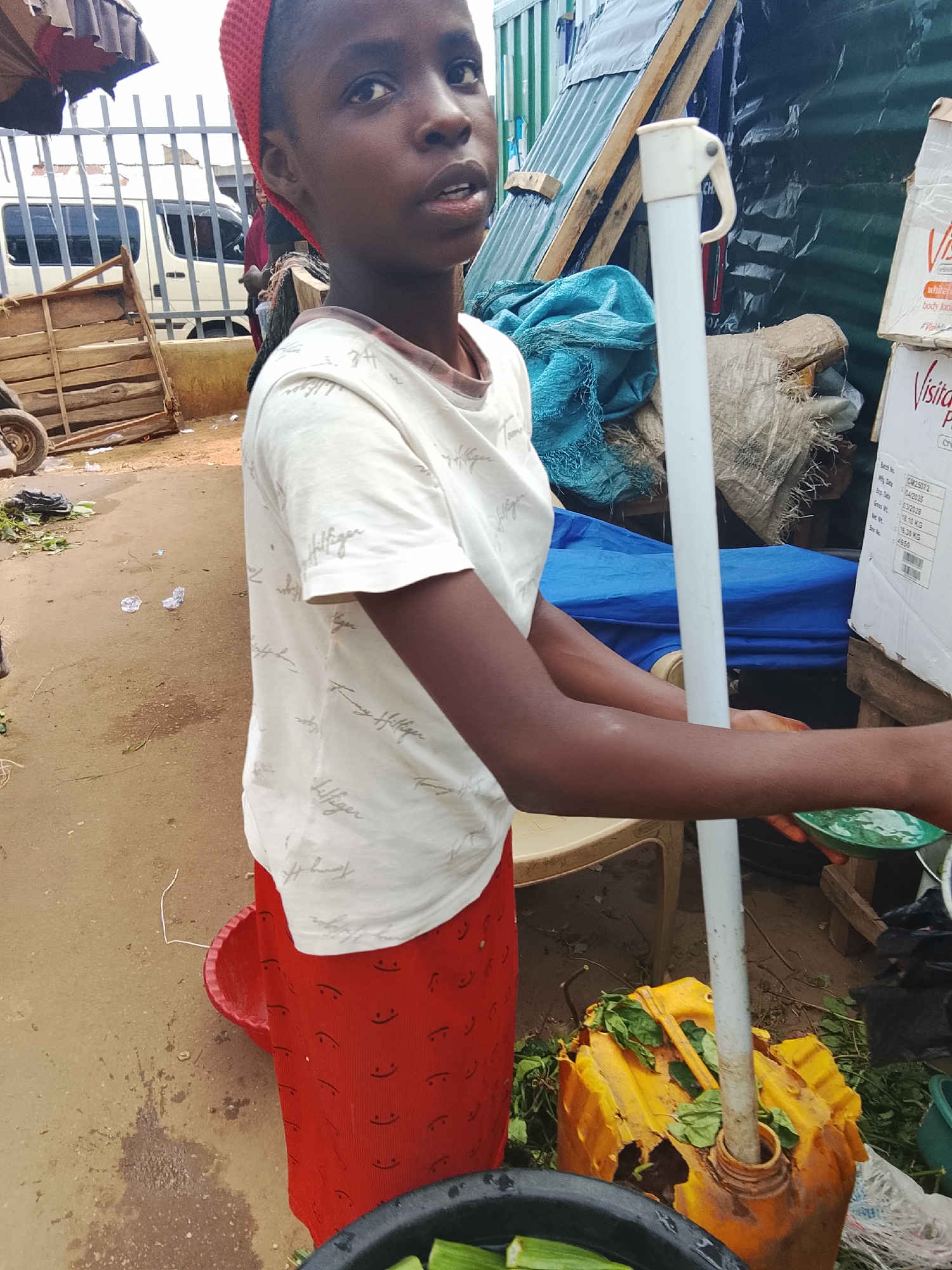 A young African girl wearing a red head covering and a white t-shirt with a repeating pattern of the "Tommy Hilfiger" logo stands in what appears to be a market. She is looking to her right with an open mouth. She is wearing a red skirt with a pattern of smiling faces. Her left arm is bent and she is holding a green plate. Her right arm is extended downwards, holding a white pipe that is inserted into a damaged yellow jerrycan. The jerrycan contains green leaves. In the background, there is a metal fence, a white van, wooden crates, and various bags and tarps. The ground is dirt and appears damp in places. The scene suggests an outdoor market environment in Akwanga, Nigeria.