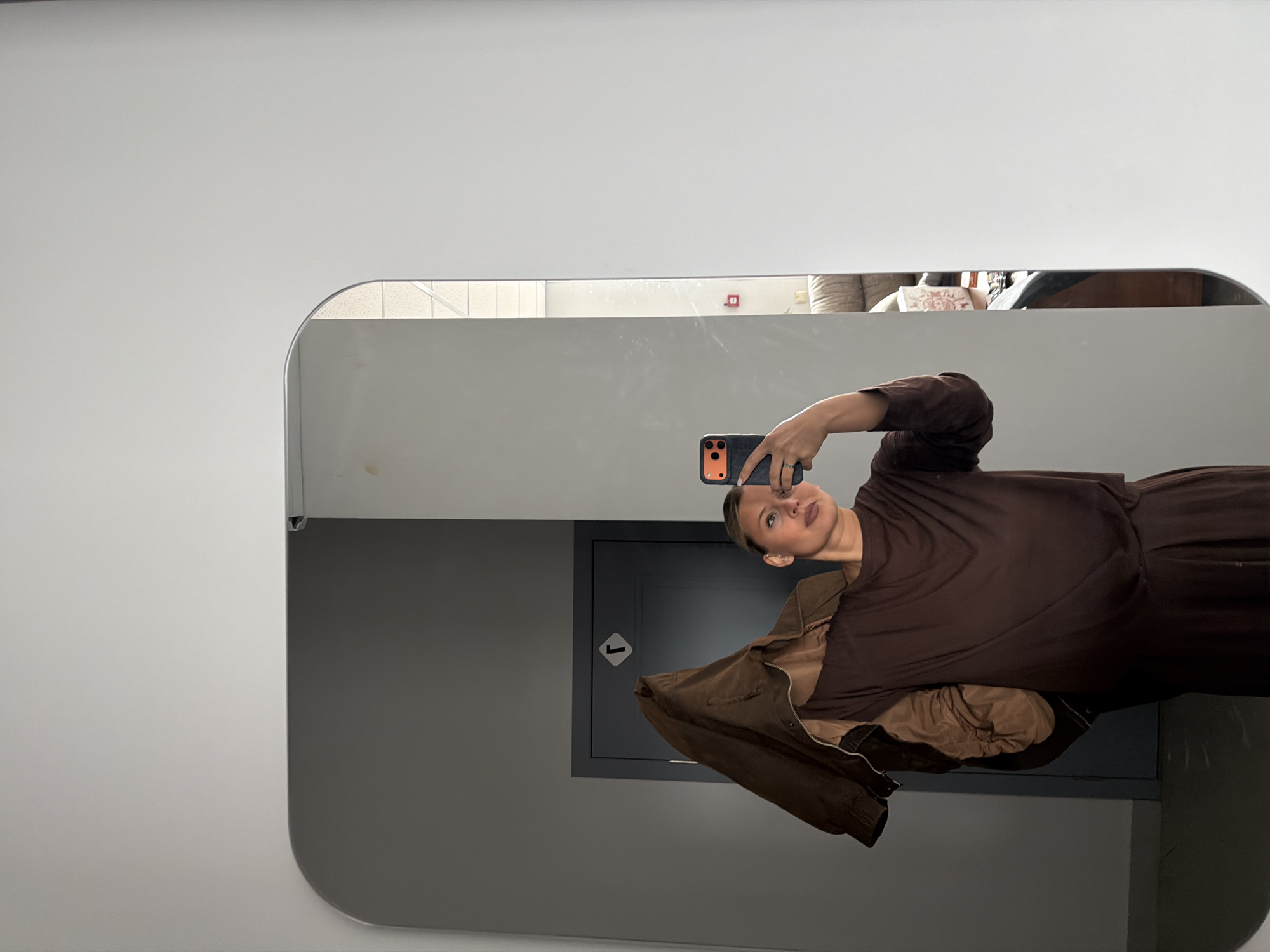 A woman is taking a selfie in a mirror. She is holding her phone in her right hand. She is wearing a brown shirt. A brown jacket hangs from her left side. The reflection shows part of an interior room, including furniture and a door with a symbol. The environment appears to be an indoor space with light-colored walls. There is no information to indicate the location is in the city of Sarapul, Russia.