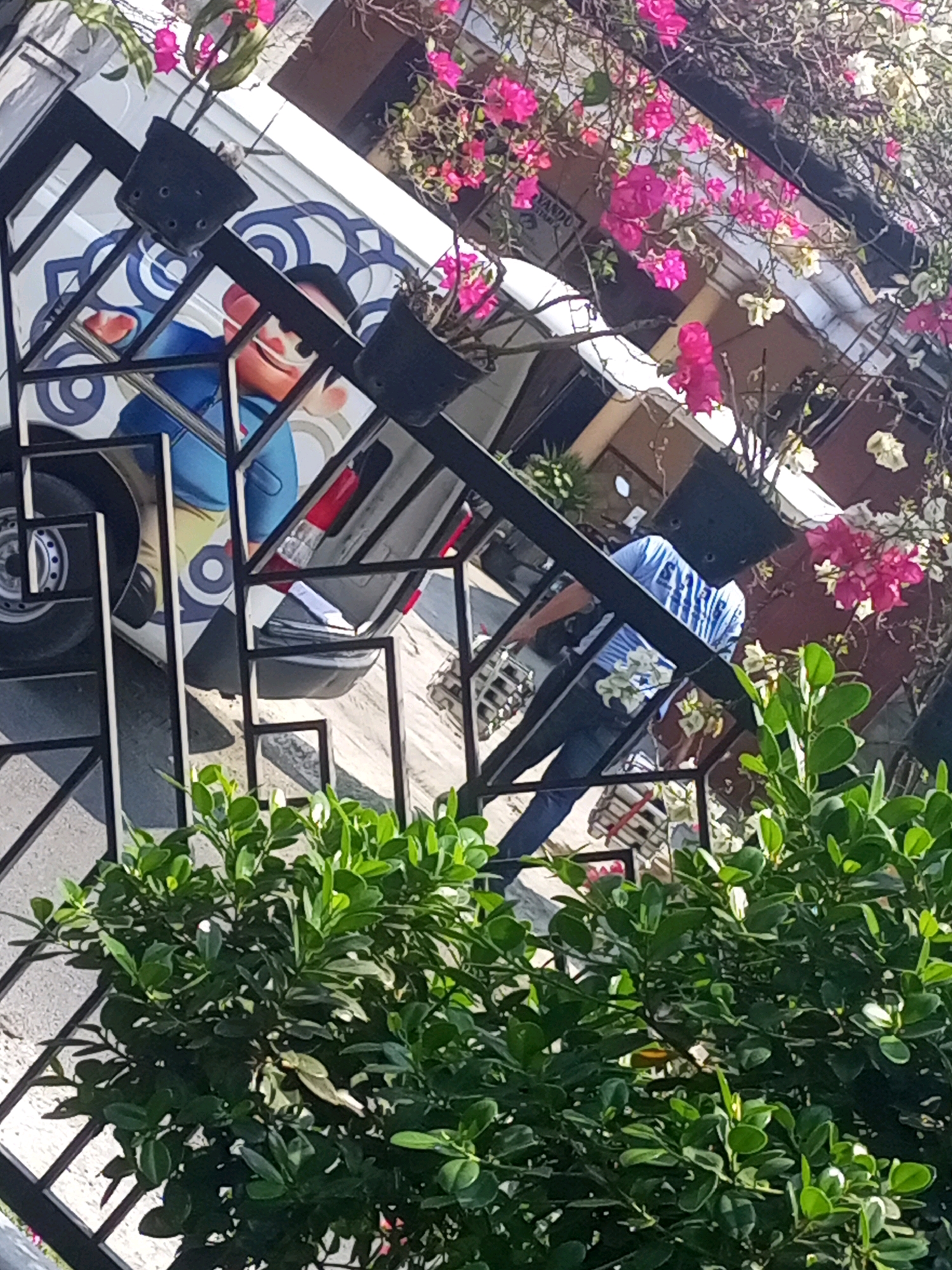 The image shows a street scene in Ngasem, Indonesia, with a focus on a white van adorned with a cartoon character. A person in a blue and white striped shirt is visible near the van, appearing to be interacting with it or its contents. The scene is framed by a black metal fence and lush green foliage, suggesting an outdoor environment. Pink bougainvillea flowers cascade from above, adding a splash of color to the composition. The perspective is slightly tilted, giving the impression of looking up at the scene.