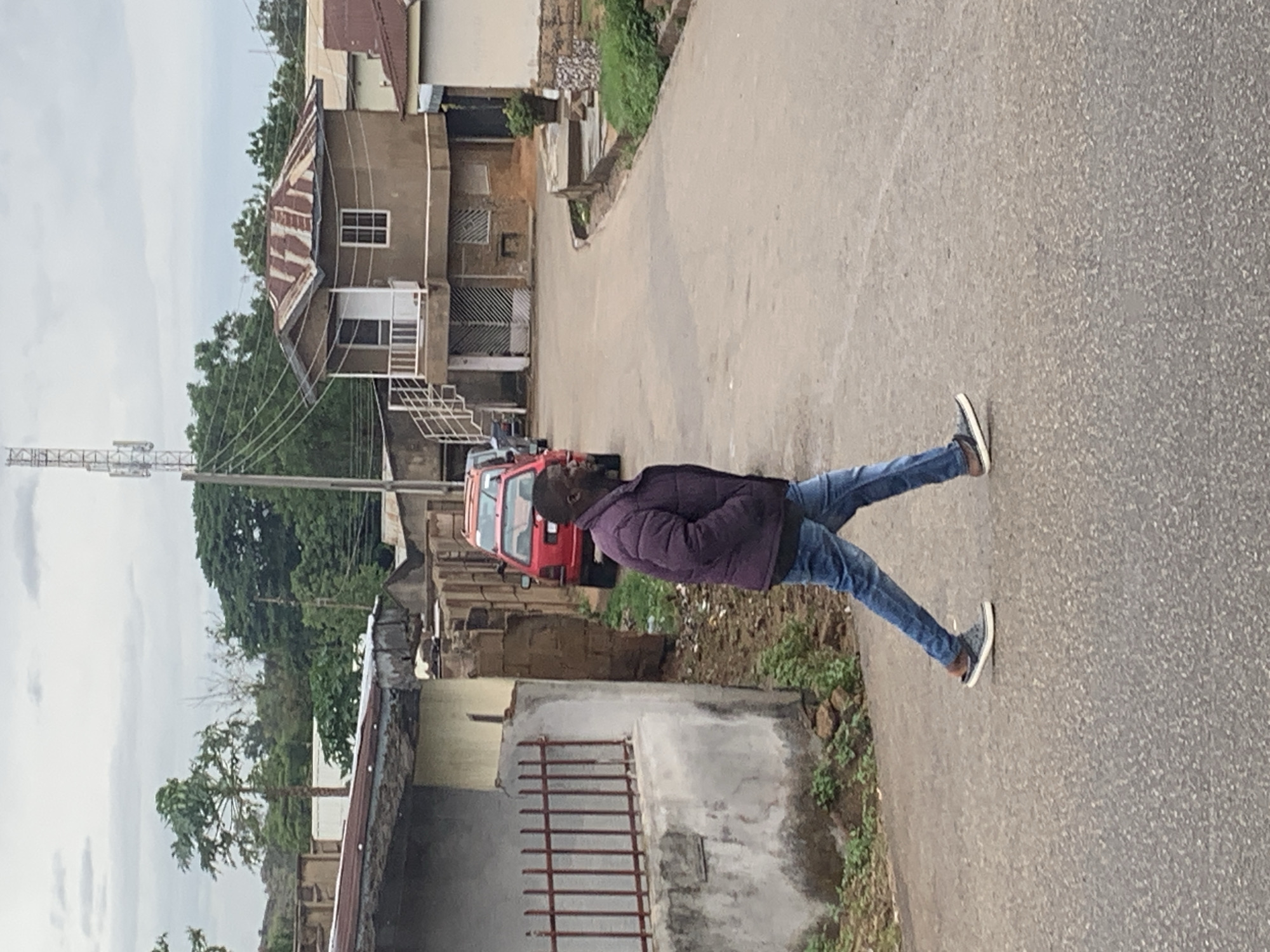 An outdoor, slightly overcast daytime scene in what appears to be a residential or semi-urban area of Jos, Nigeria. A man stands on a paved road, facing slightly away from the camera. He is wearing a dark purple or maroon puffer jacket, blue jeans, and dark sneakers with white soles. His stance is casual, with one leg slightly bent.
Behind the man, a red car is parked partially visible on the side of the road. The background features several buildings of varying architectural styles and conditions. One prominent building has a rusty, corrugated metal roof, light-colored walls, and a white-framed window, with a white staircase or railing leading to an upper level. Further in the background, a tall, slender metal telecommunication mast stands against the cloudy sky. Trees and sparse green vegetation are scattered throughout the scene, including a papaya tree. Utility wires crisscross overhead. The road itself is gray asphalt. No specific activity beyond the man standing is occurring, and no discernible text is visible anywhere in the image.