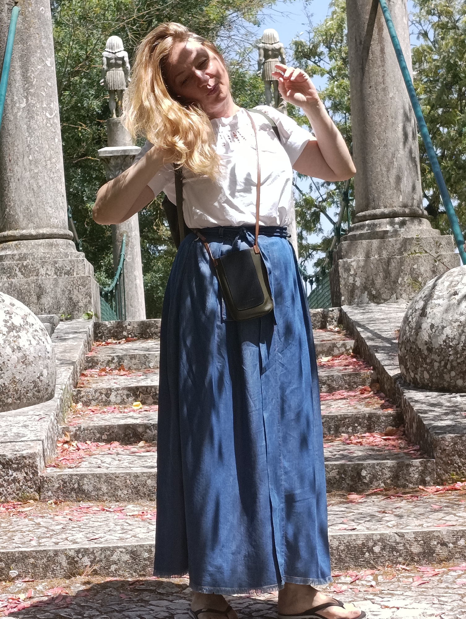 The image shows a woman standing on stone steps outdoors. She has long blonde hair and is wearing a white top and a long blue denim skirt. A dark green crossbody bag hangs from her shoulder. The steps are lined with rose petals and appear to be in a garden or park setting. In the background, there are large stone pillars, trees, and a glimpse of a blue sky. The architectural style and the presence of stone sculptures on pedestals suggest a historical or monumental location. This location is the Palácio da Pena in Sintra, a town near Lisbon, Portugal.