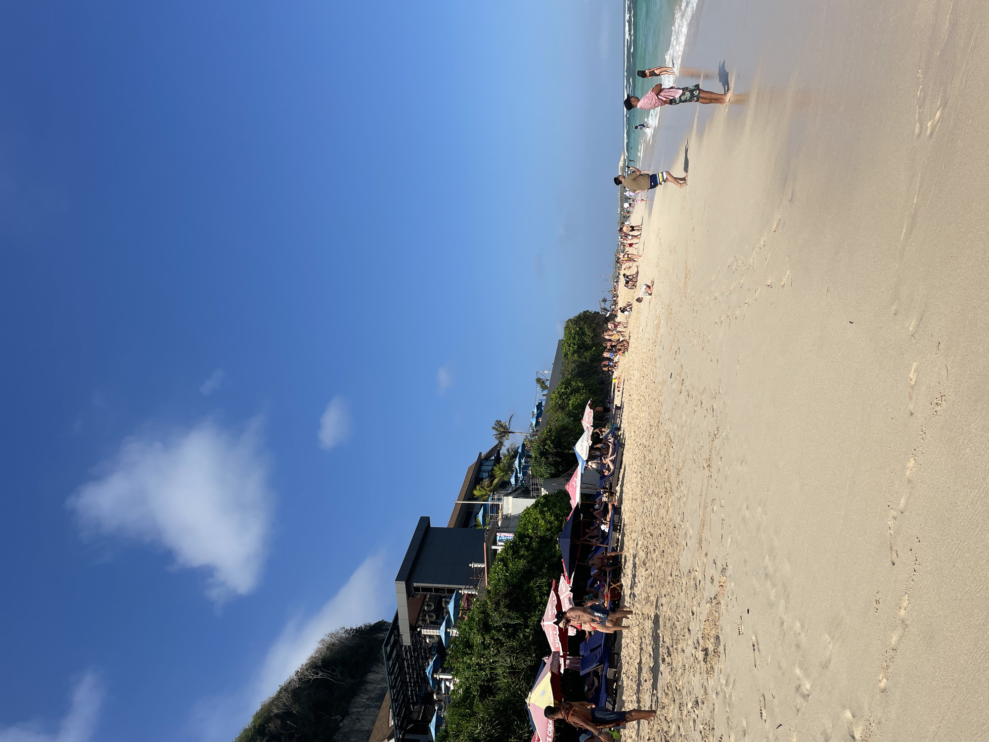 The image depicts a bustling beach scene in Kuta Selatan, Indonesia, on a sunny day. The foreground is dominated by fine, light-colored sand marked with numerous footprints. In the midground, a stretch of beach is lined with colorful umbrellas, lounge chairs, and several groups of people. Some individuals are walking along the shore, while others are seated or reclining on the chairs. Further back, a modern building complex, possibly a resort or restaurant, is situated on a cliffside, adorned with blue umbrellas. The ocean occupies the right side of the frame, with clear turquoise water and small waves. The sky is a vibrant blue with a few scattered white clouds. The overall atmosphere suggests a popular tourist destination.