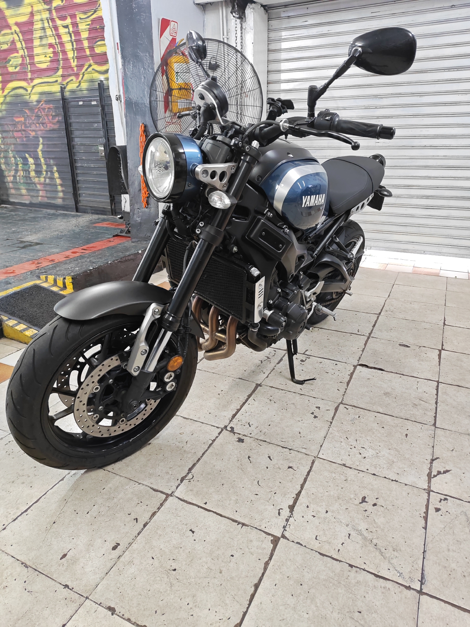 The image shows a Yamaha XSR900 motorcycle parked on a tiled surface, likely a sidewalk or garage floor. The motorcycle is primarily black and blue with silver accents and has a circular headlight. The background includes a closed metal shutter, a partially visible fan, and a wall with graffiti.
It is impossible to verify the location of this image or its subject in the city of Buenos Aires, Argentina.