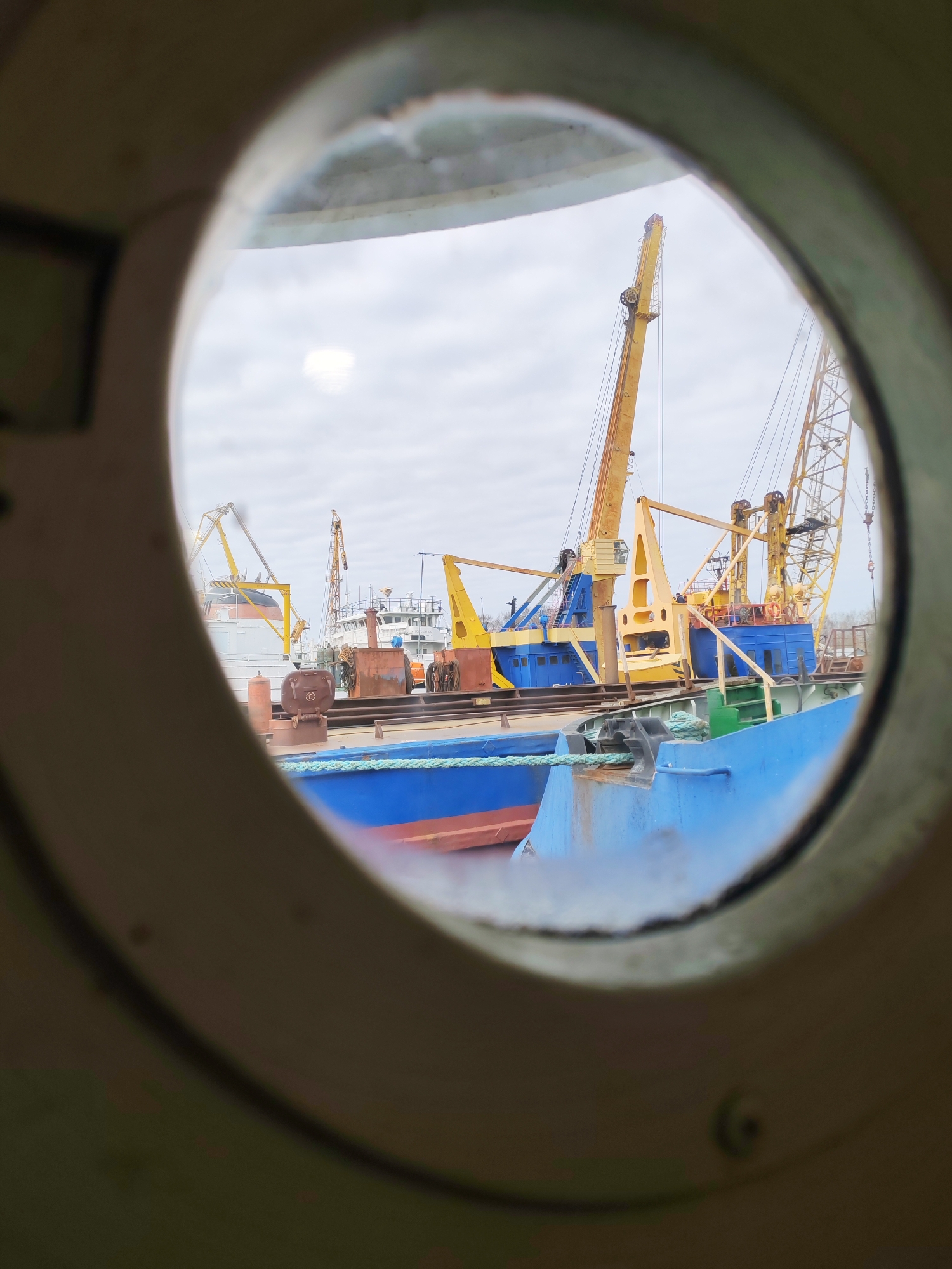 The image provides a detailed view of a bustling port or shipyard, framed by the circular opening of a ship's porthole. The porthole itself is metallic, possibly olive green or gray, and appears slightly worn or smudged, with a prominent bright reflection on its glass in the upper left quadrant.
Through the porthole, multiple large vessels and industrial equipment are visible under an overcast daytime sky. In the foreground and mid-ground, a blue-hulled ship is prominently docked, secured by a thick, braided green-blue mooring rope tied to a dark bollard. Its deck features various metallic structures, some appearing rusty brown, including what looks like a hatch or container lid, and one structure bears the faint numbering "02".
Behind this vessel, another large ship, painted in distinct blue and yellow sections, is central to the scene. This ship is equipped with several towering yellow cranes, suggesting active cargo loading, unloading, or maintenance operations. On one of these yellow crane structures, faint Cyrillic text, possibly "ЧОМТП", is discernible. In the background, additional ships with white superstructures and yellow cranes further emphasize the industrial nature of the setting. No people are visible in the scene. The overall impression is one of maritime industry and activity under a bright but cloudy sky.