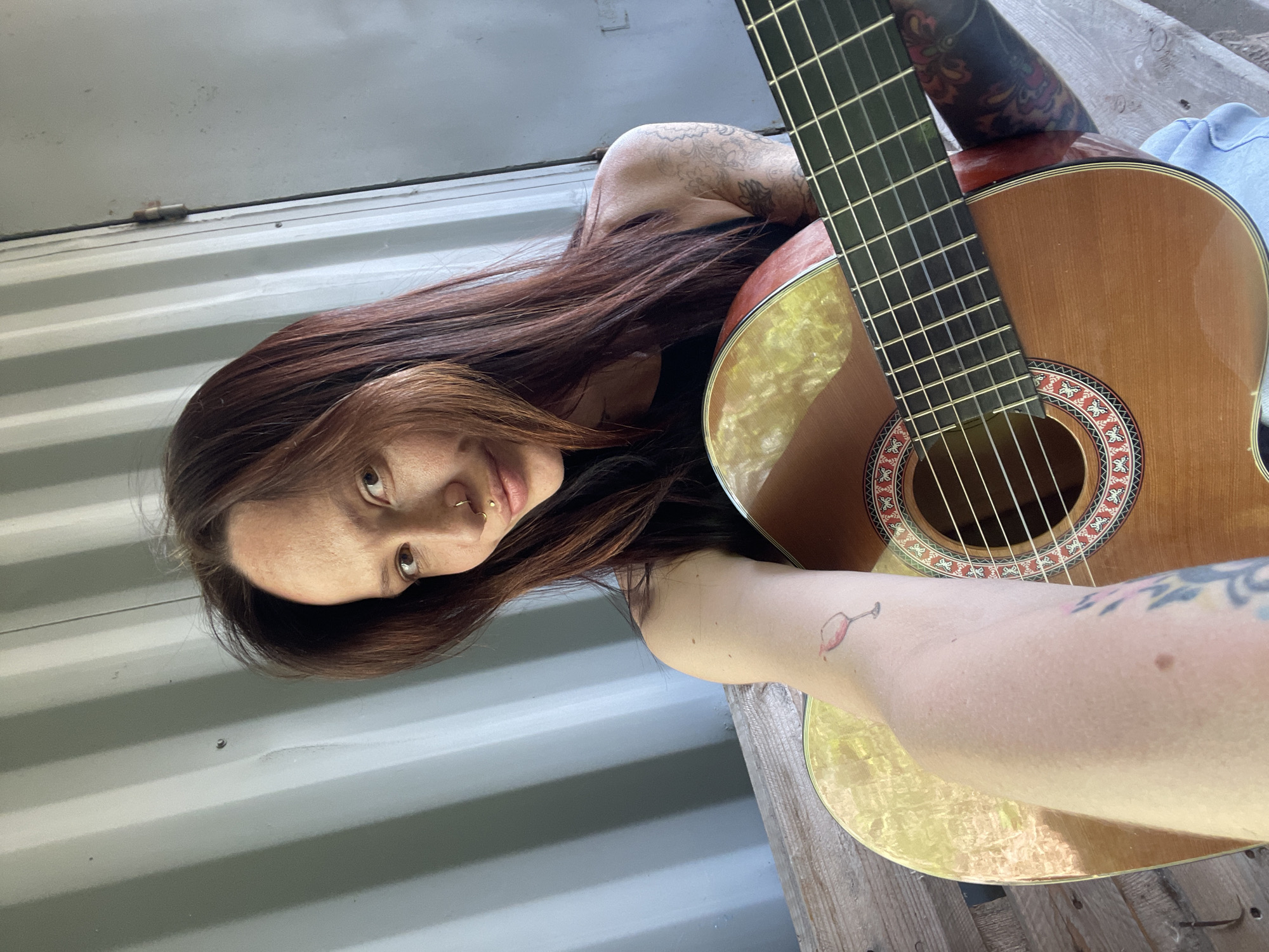 A woman with long, auburn hair, a lip piercing, and tattoos on her shoulder and arm is lying on what appears to be a wooden surface next to a corrugated metal wall. She holds an acoustic guitar with her tattooed arm. The location is consistent with a rural or semi-industrial setting. There is no definitive evidence to confirm this photograph was taken in the city of Kabanikha, Russia.