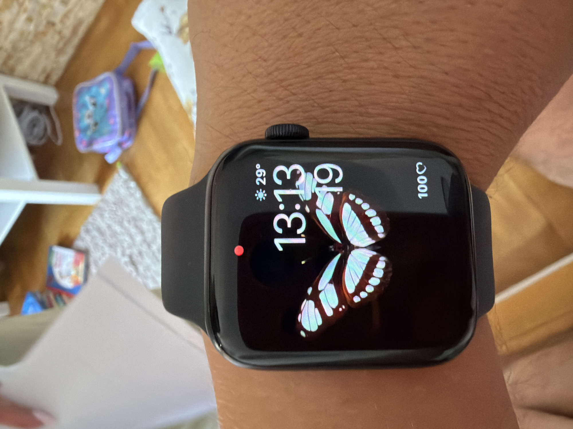 A close-up shot shows a black Apple Watch on a person's wrist. The watch face displays a digital time of "13:13" and the date "19". A graphic of a butterfly with blue and white wings is featured on the watch face. To the upper right of the time, the temperature is shown as "29°" with a sun icon. At the bottom right of the watch face, "100♡" is displayed. The background is blurred, but appears to be an indoor setting with wooden flooring, a rug, and a white shelf with items on it, including what looks like a purple backpack. The scene is not in Budva, Montenegro.