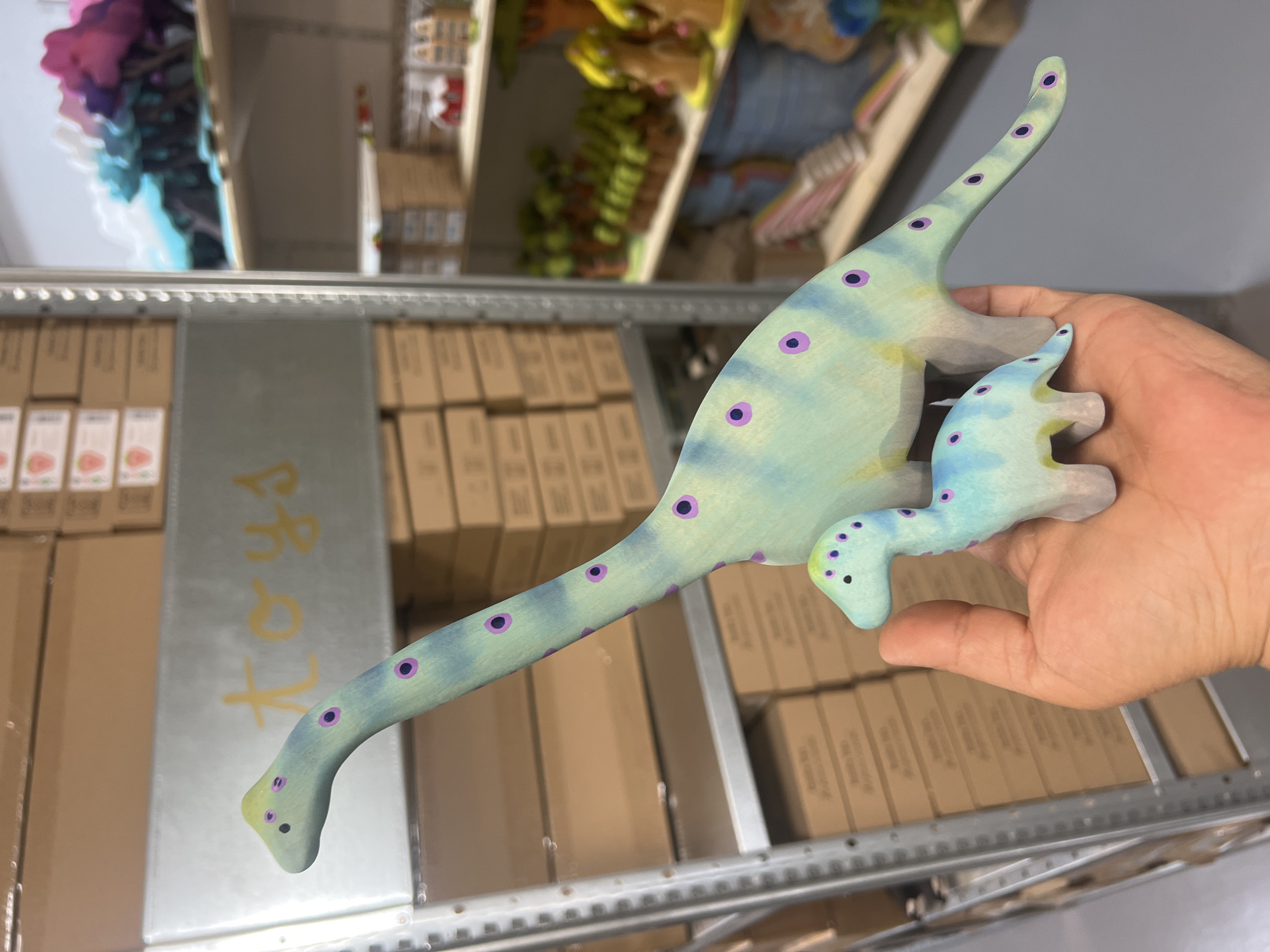 Two wooden toy dinosaurs, one significantly larger than the other, are displayed in a person's palm. The larger dinosaur is a pale green with purple spots and a yellow underside, and the smaller dinosaur is a lighter blue-green with similar purple spots. They are held in front of shelves stacked with cardboard boxes in what appears to be a warehouse or storeroom. The background shelves also contain other colorful toys, including what look like wooden figurines and colorful trees. A sign with the word "TOYS" written in yellow script is visible on a shelf behind the dinosaurs. The lighting suggests an indoor environment with overhead or diffused lighting.