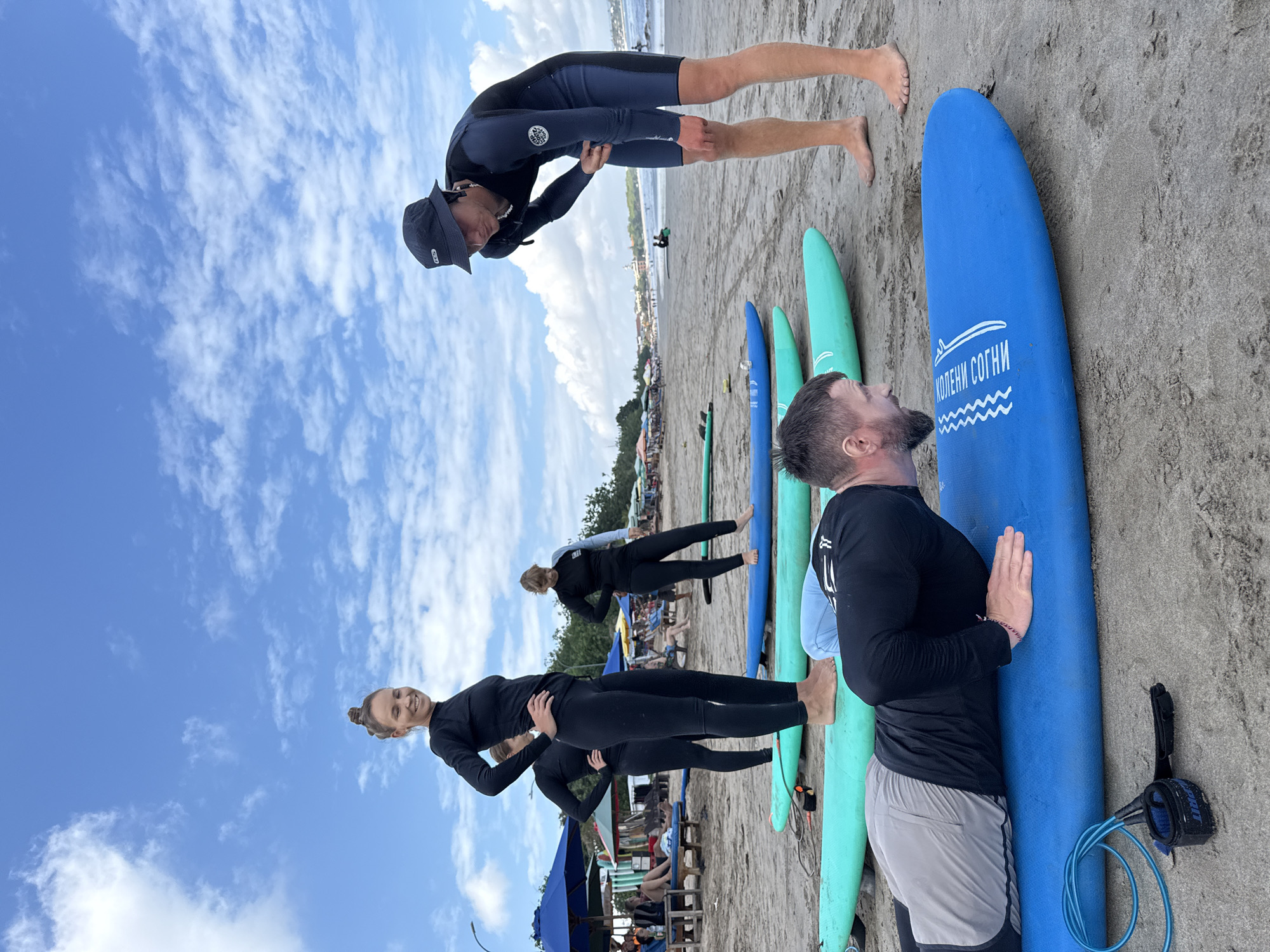 A group of people are on a sandy beach in Kuta, Indonesia, gathered around several surfboards. In the foreground, a man with a beard is lying face down on a blue surfboard, his hands positioned as if preparing for a push-up. He is wearing a black long-sleeved rash guard and grey shorts. To his right, a young man wearing a dark blue and black wetsuit and a dark bucket hat is bending over, looking down at the surfboard. Behind them, three women in black wetsuits are standing or posing on surfboards. The woman on the far left is smiling and looking towards the camera, with her hands on her hips. The middle woman is standing with her hands on her hips, and the woman on the far right is looking down. In the background, more surfboards are lined up, and beach umbrellas and lounge chairs are visible, along with people relaxing. The ocean and a cloudy blue sky complete the scene.
