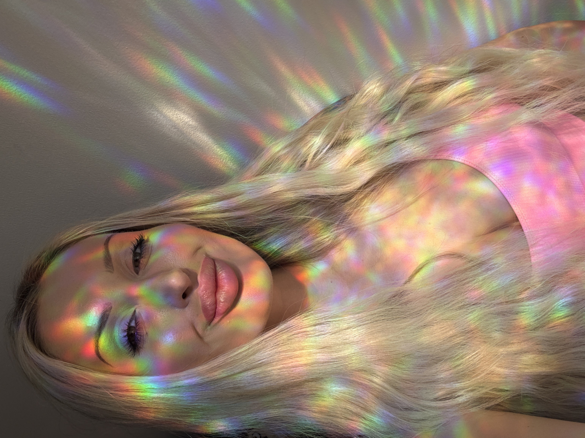 A close-up portrait shows a fair-skinned woman with long, blonde hair. She is looking at the camera with a slight smile and her left eye is partially closed. Her face is illuminated by a spectrum of rainbow-colored light, casting colorful patterns across her skin, hair, and a pink top. The light appears to be coming from a prism or a similar light-refracting object, creating a playful and ethereal effect. The background is a textured wall, also illuminated by the colorful light. The lighting suggests an indoor setting, potentially near a window.