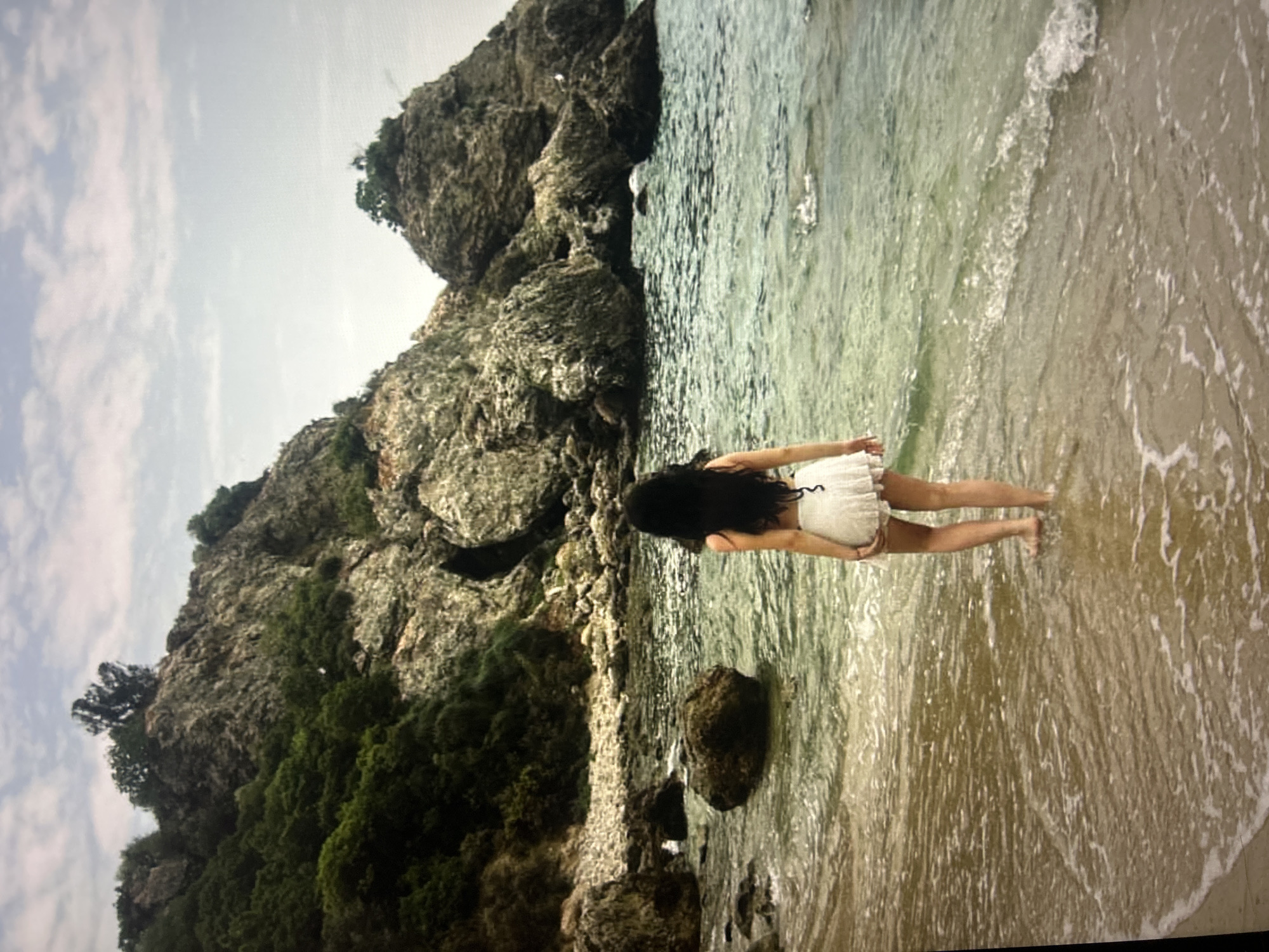 A woman in a white dress stands in shallow water at a beach with a rocky coastline. The beach extends from the lower right, with clear, light-colored water washing onto the sand. She is facing away from the camera, with her dark hair flowing down her back. The coastline consists of large rocks and lush green vegetation. The sky is partially cloudy. The location is unknown but there is no obvious visual information to support that it is in Sofia, Bulgaria.