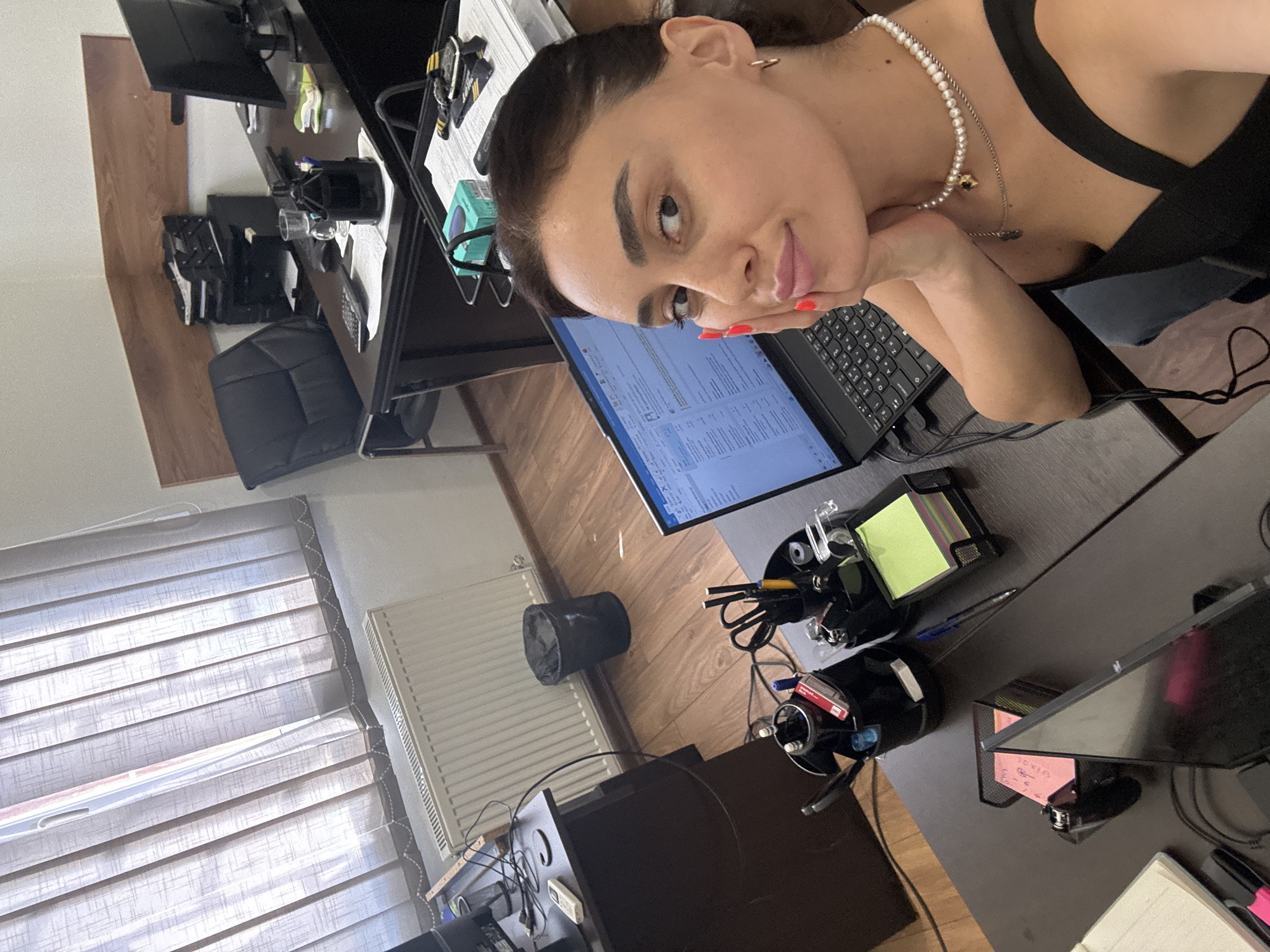 A woman is taking a selfie in an office in Tbilisi, Georgia. She is sitting at a desk with a laptop in front of her. She is wearing a black top and a pearl necklace. The office is cluttered with papers, pens, and other office supplies. There is a window with blinds in the background. The woman is looking at the camera with a slight smile.